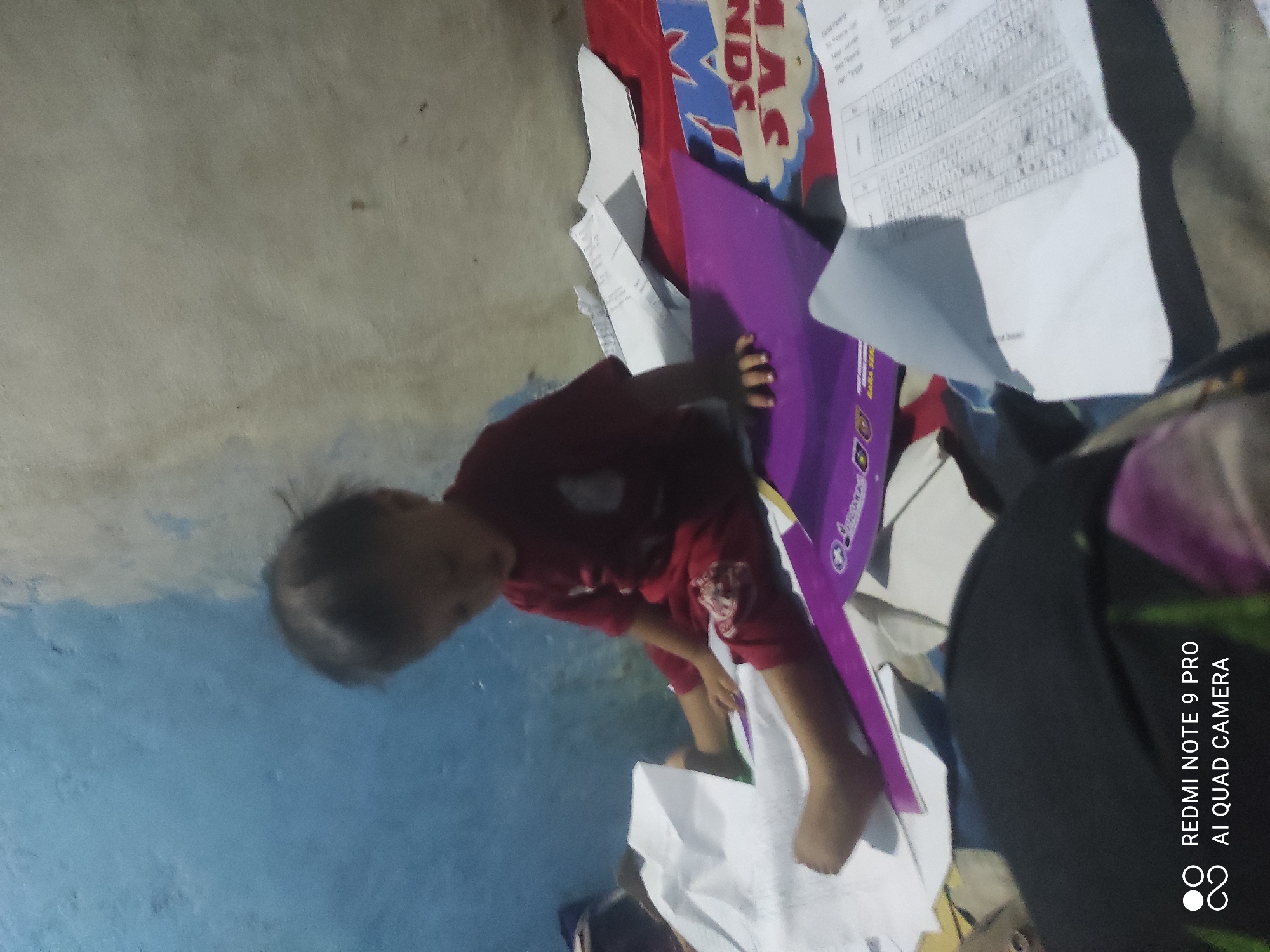 A child is sitting on a bed, surrounded by papers and a purple folder. The child is wearing a red t-shirt and red shorts. The room appears to be a bedroom, with a blue and beige painted wall in the background. There are also some sheets of paper scattered around the child. The bottom left corner of the image contains a watermark indicating "REDMI NOTE 9 PRO AI QUAD CAMERA." The context of Sakra, Indonesia is not visually discernible from the image.