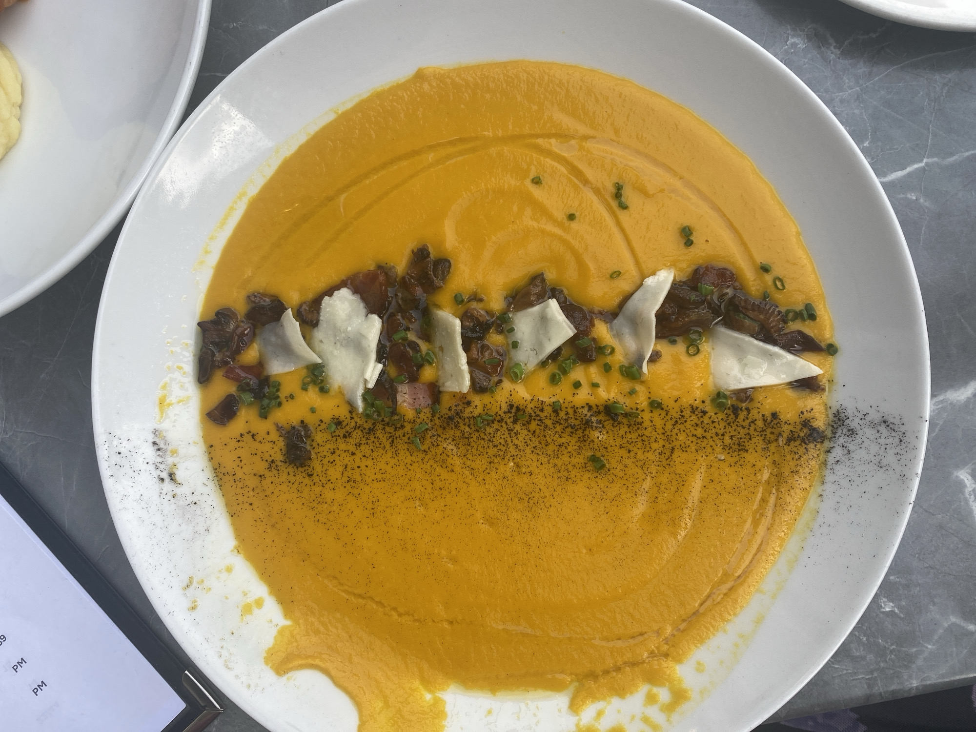 An overhead shot shows a white oval bowl filled with bright orange soup. The soup is garnished with a line of crumbled dark brown ingredients, thin white shavings, and finely chopped green herbs. A dusting of black powder, possibly pepper or ash, covers the left side of the soup. The bowl sits on a grey marble surface. In the upper left corner, a portion of a menu with text is visible. The scene appears to be indoors, possibly in a restaurant, with no people visible. The setting is not specified to be in Montreal, Canada.