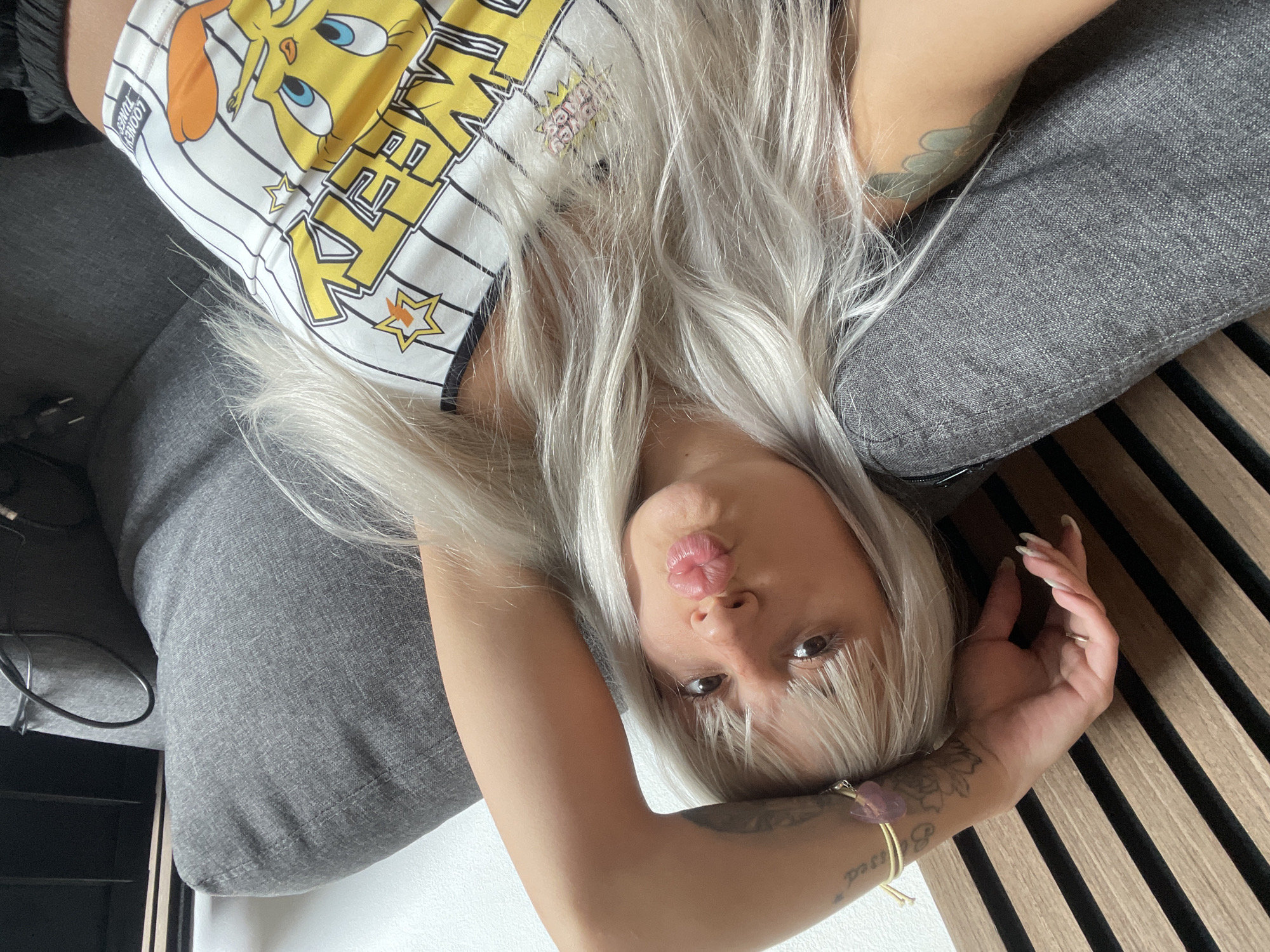 A young woman lies on a grey sofa in a room with wooden slat detailing in the background. She is upside down relative to the camera, wearing a yellow and white striped tank top with a cartoon character on it. She has long, platinum blonde hair with bangs, and is pouting with her tongue slightly visible. Her left arm is bent and raised near her face, showing tattoos and bracelets on her wrist. The setting does not suggest any known location in Tilburg.
