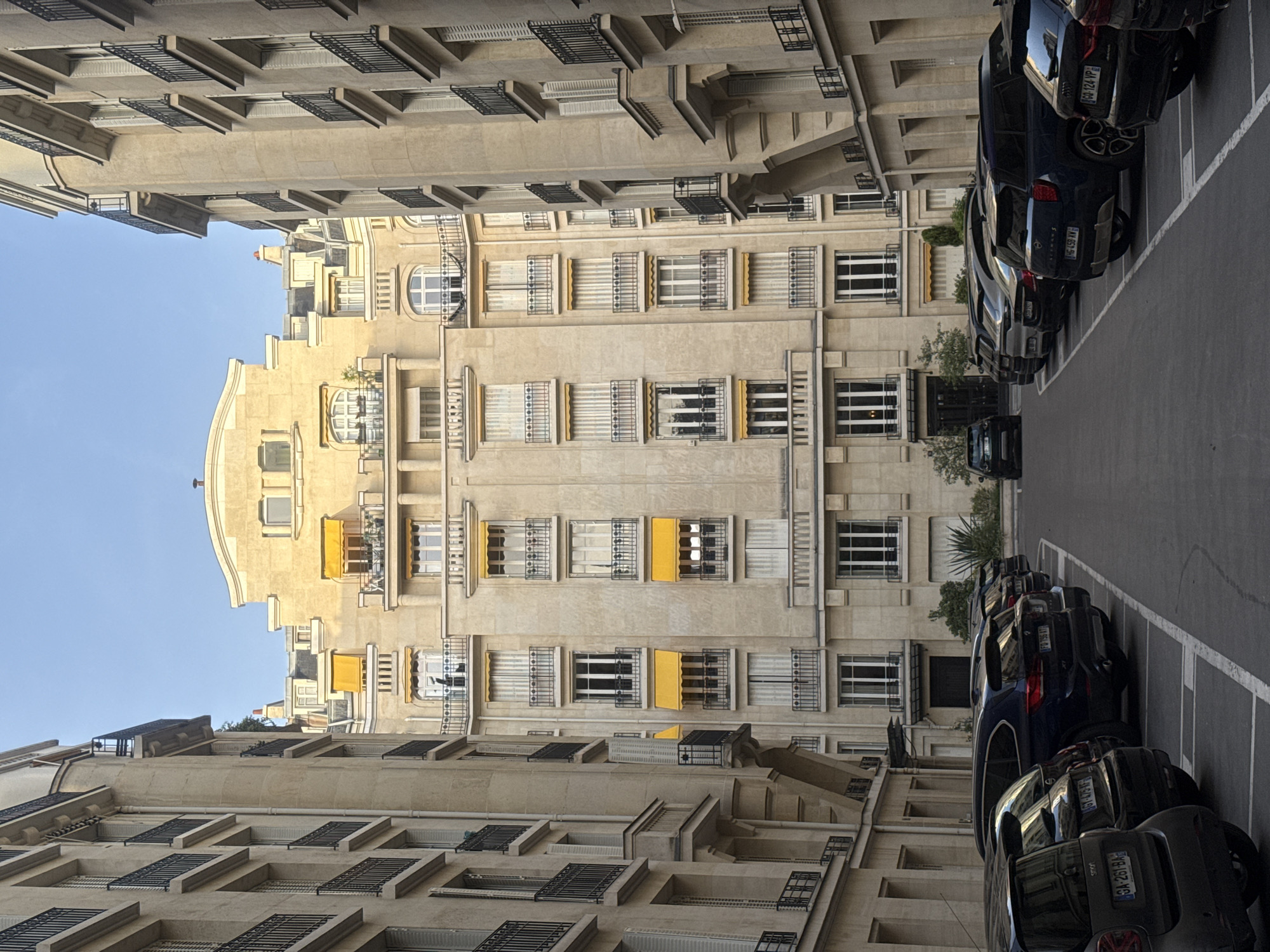 The image shows a street in Paris, France, lined with tall, light-colored buildings. The architecture of the buildings is ornate, with many windows, balconies, and decorative details. Some windows have yellow awnings. Cars are parked along the side of the street. The sky is visible in the upper left corner, and it appears to be a clear day. License plates are visible on the cars, with identifiers such as "GA-261-BA", "GT-475-IT", "HG-458 XV", and "GQ-124-VP". The image is taken at street level.