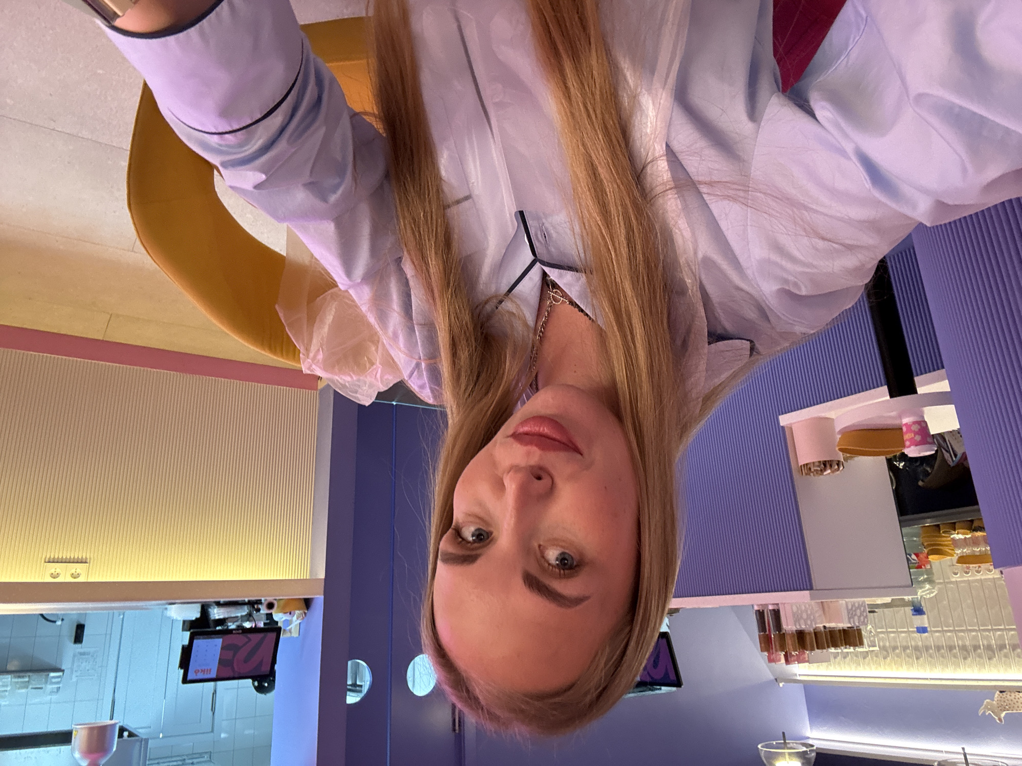 A fair-skinned woman with long blonde hair, wearing a light blue pajama-style top with black piping, is taking a selfie. She is indoors, possibly in a cafe or restaurant, as suggested by the background: a yellow chair, purple ribbed walls, and a counter with food displays. The lighting is bright and artificial. A reflective surface creates a mirror image of the counter and displays. The presence of a screen suggests a point-of-sale system. The location is Sochi, Russia.