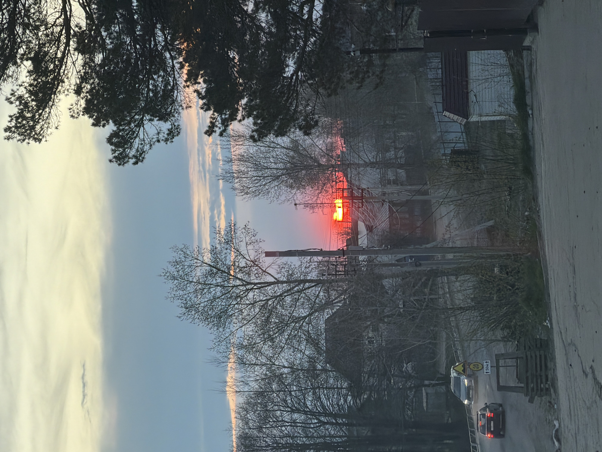 The image depicts a street scene in Chaikovsky, Russia, possibly during sunset or sunrise. The sky is visible above buildings and trees, with the sun appearing as a bright red/orange glow. Silhouette trees, telephone poles, and buildings are partially visible. In the lower right foreground, a road with two vehicles is present, one being a light-colored vehicle and the other being a dark-colored vehicle. Road signage is also observable near the light-colored vehicle. The image is oriented vertically.