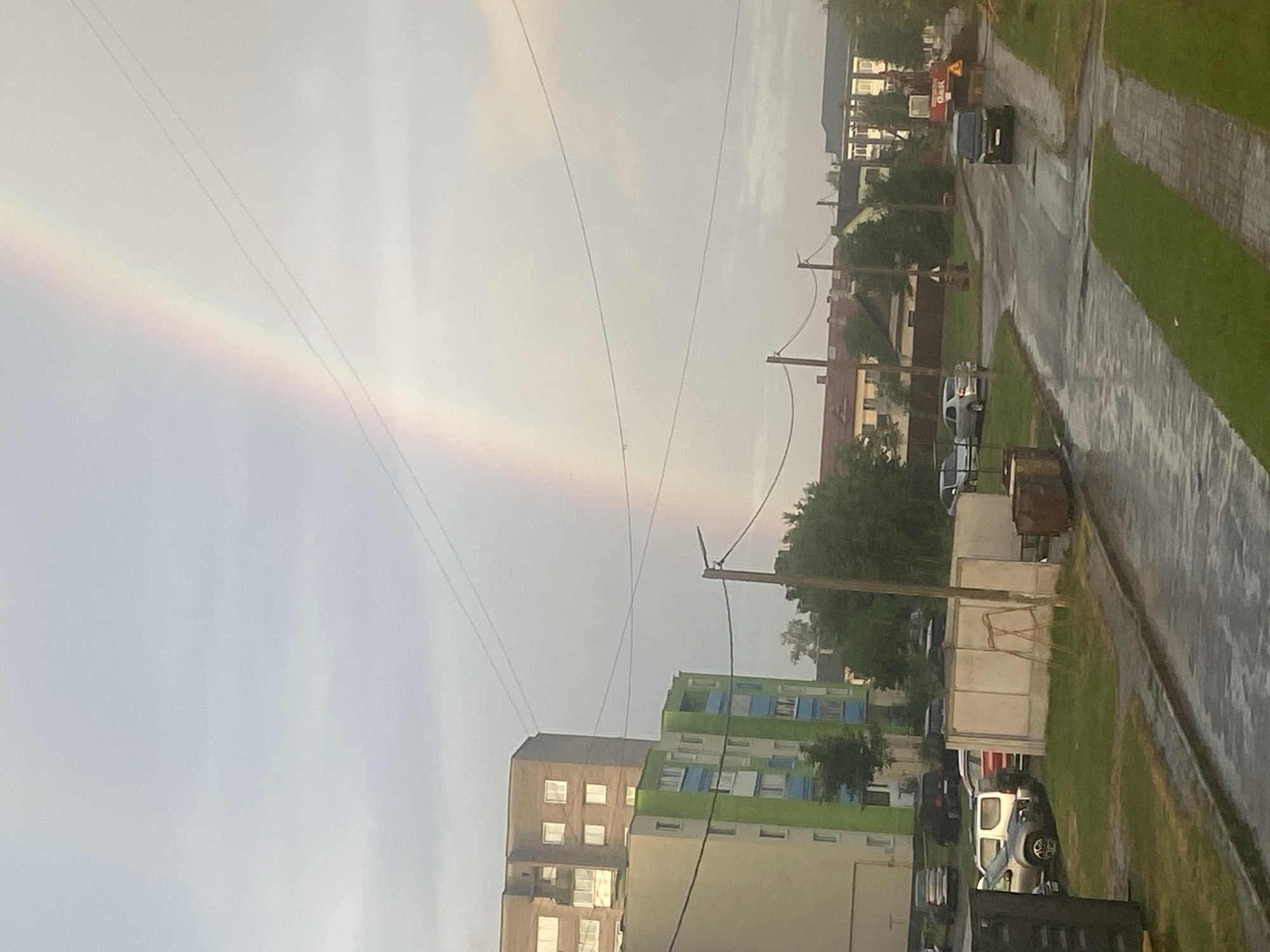 The image depicts a street scene in Krupki, Belarus, under a cloudy sky where a faint rainbow is visible. In the foreground, a wet asphalt road curves to the right, bordered by a grassy median strip with paving stones. To the left, several parked cars, including a silver SUV and a dark sedan, are visible near a large, neutral-colored temporary structure or wall. Adjacent to this structure is a trash bin. Further left, a light-colored building with balconies and windows is partially visible. In the background, two residential buildings stand tall; one is taller and brown, the other is shorter and painted green and light yellow. Trees are scattered throughout the scene. Across the road, smaller buildings with reddish-brown roofs are present. A black van is driving on the road, and a construction vehicle, possibly an excavator, with the word "GOMEL" visible on its side, is operating on the right side of the road. Power lines crisscross the sky.