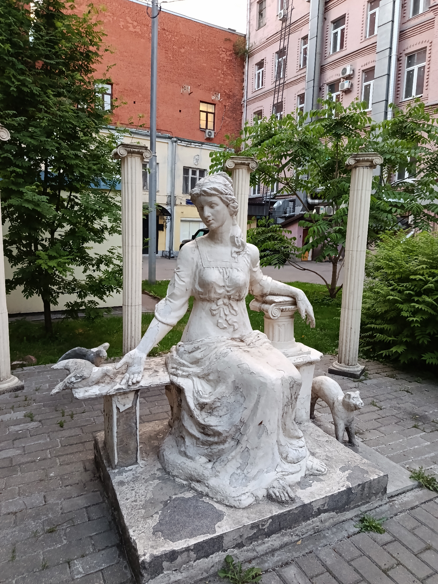 The image shows a sculpture of a seated woman in a classical style, situated outdoors. The sculpture is rendered in white and appears aged or distressed, with visible cracks and wear. She is depicted wearing a long dress and her hair is styled in a loose braid. Her right arm rests on a small column while her left hand is near a sculpture of a cat and several birds to her left. Another cat is at her right.
The sculpture is positioned in a courtyard or garden setting with brick pavement and several columns with Corinthian capitals behind and around the statue. Behind the columns is foliage from trees and shrubs. Buildings are visible in the background, constructed of brick and painted in different colors, including a red brick building and a light pink building. A utility pole is also visible.
Based on the style of architecture and context, this image likely originates from Moscow, Russia.