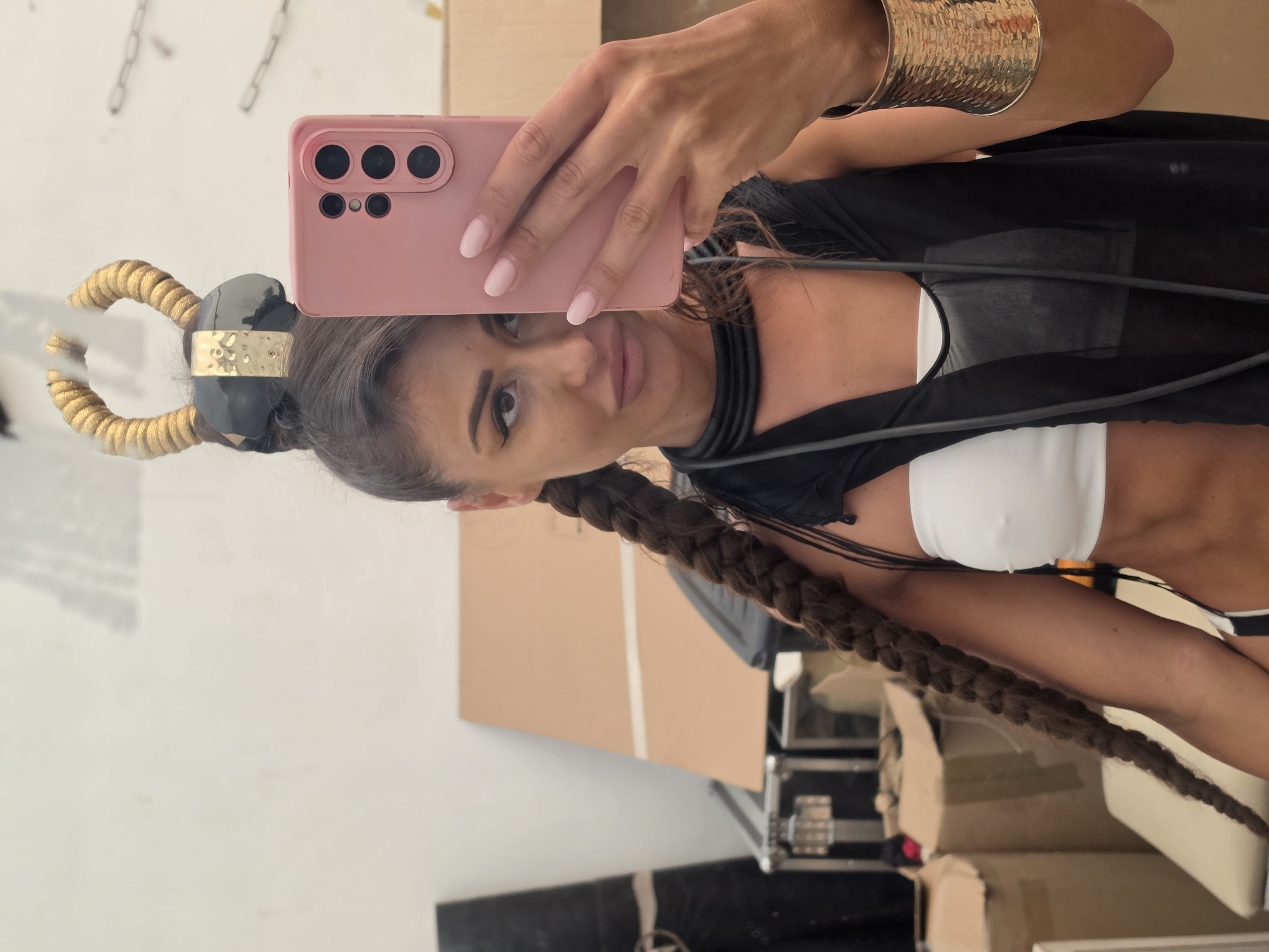 A woman takes a selfie in a room with a white wall and cardboard boxes in the background. She has dark hair pulled back with a gold and black accessory on top of her head and a long braid. She is wearing a white bandeau top and a sheer black overlay. She is also wearing a black choker necklace and a wide gold bracelet. She is holding a pink smartphone in her right hand. The room appears to be an indoor storage area or backstage.