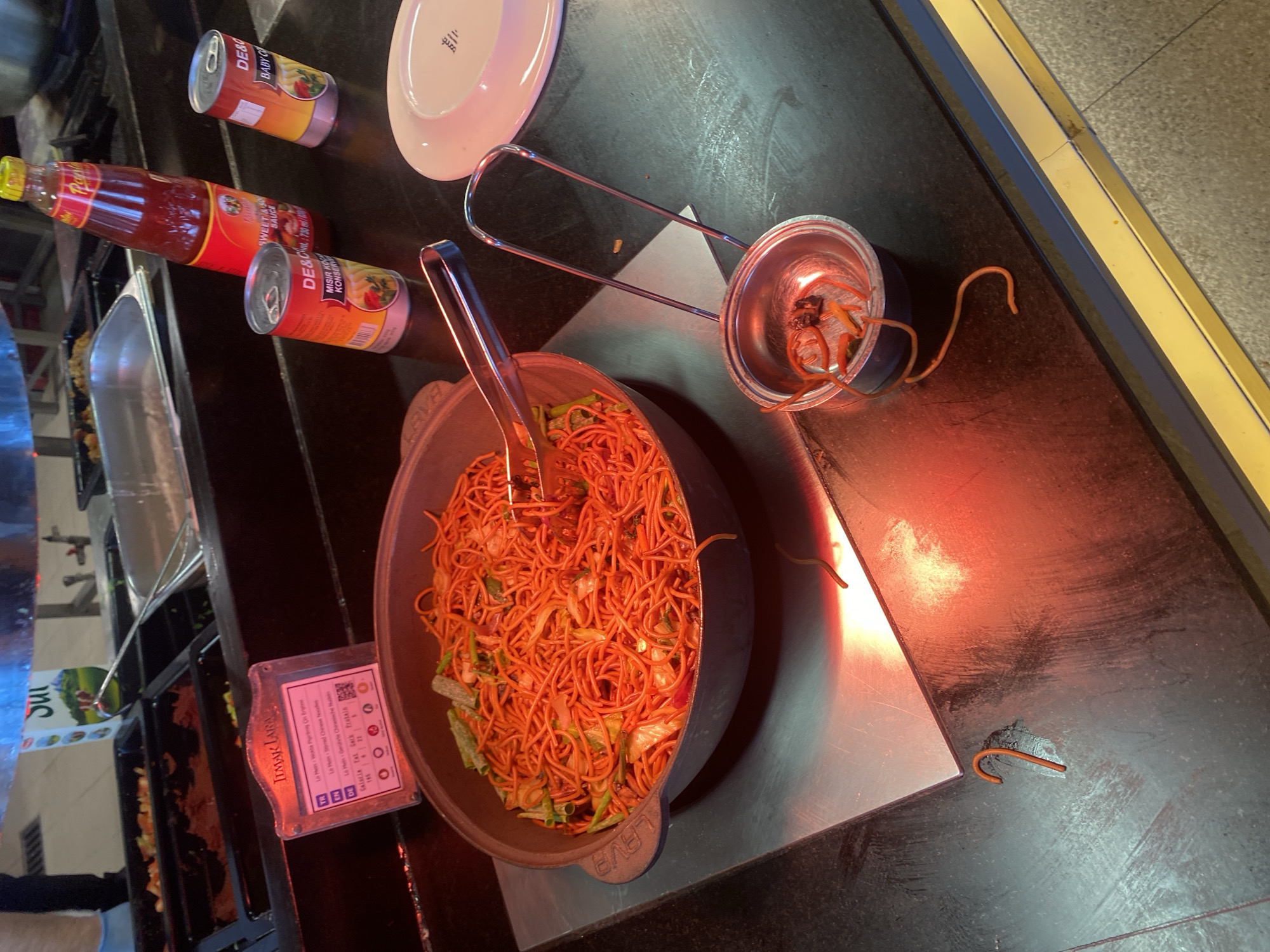 The image shows a buffet-style food service counter, likely in Aksu, Turkey, given the presence of Turkish text on a food label. In the foreground, a large dark bowl filled with red-orange noodles and chopped green vegetables is prominently displayed. Tongs rest inside the bowl, suggesting recent serving. To the right, a small metal ladle sits on the counter, containing a few strands of noodles and some dark specks, possibly food debris. Further to the right, a white plate is partially visible. Behind the noodles, a red bottle of sauce and two small silver cans are arranged on the counter. The scene is illuminated by warm lighting, creating a reddish glow on the surfaces. A metal serving tray with food can be seen in the background. A sign with text in Turkish, English, and German is positioned to the left of the noodle bowl, indicating details about the dish.