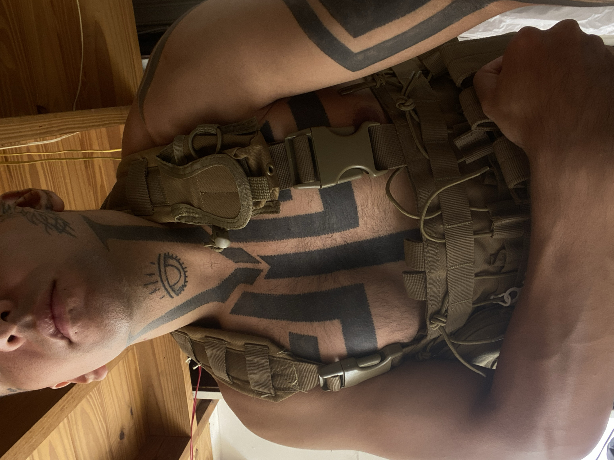 A medium-shot photograph presents a light-skinned person wearing a tan tactical vest. The subject has prominent black tribal-style tattoos covering their upper body, including the neck, chest, and upper arms. A distinctive tattoo resembling an eye is visible on the left temple. The tactical vest appears to be constructed from nylon webbing and features multiple straps and buckles. The person's face is partially visible, with their lips slightly parted. The background includes a wooden structure and some wires, offering a glimpse of the indoor environment. There are no indications in the image that point to the location in the city of Buenos Aires, Argentina.