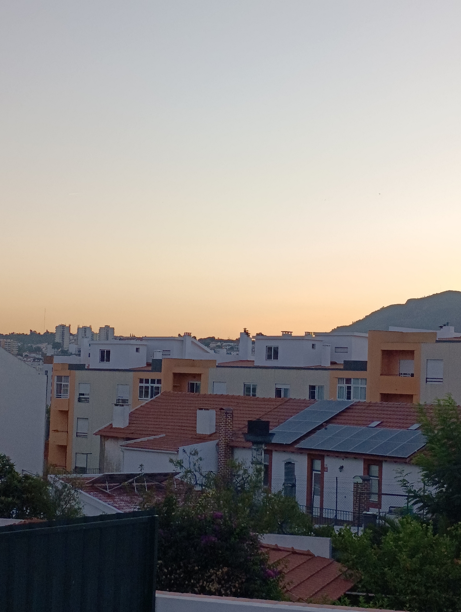 The image depicts a cityscape at dusk or dawn, with buildings of various heights and architectural styles. The sky is a gradient of pale orange and yellow, transitioning to a lighter blue towards the top. In the foreground, there are several residential buildings with red tile roofs. Some of these buildings have solar panels installed on their roofs. Further in the background, there are taller apartment buildings and a distant hillside. The scene appears to be in a suburban or urban area, likely Setúbal, Portugal, given the characteristic building designs and the presence of hills. There are no people visible in the image. The overall impression is one of a tranquil, residential environment bathed in soft, natural light.