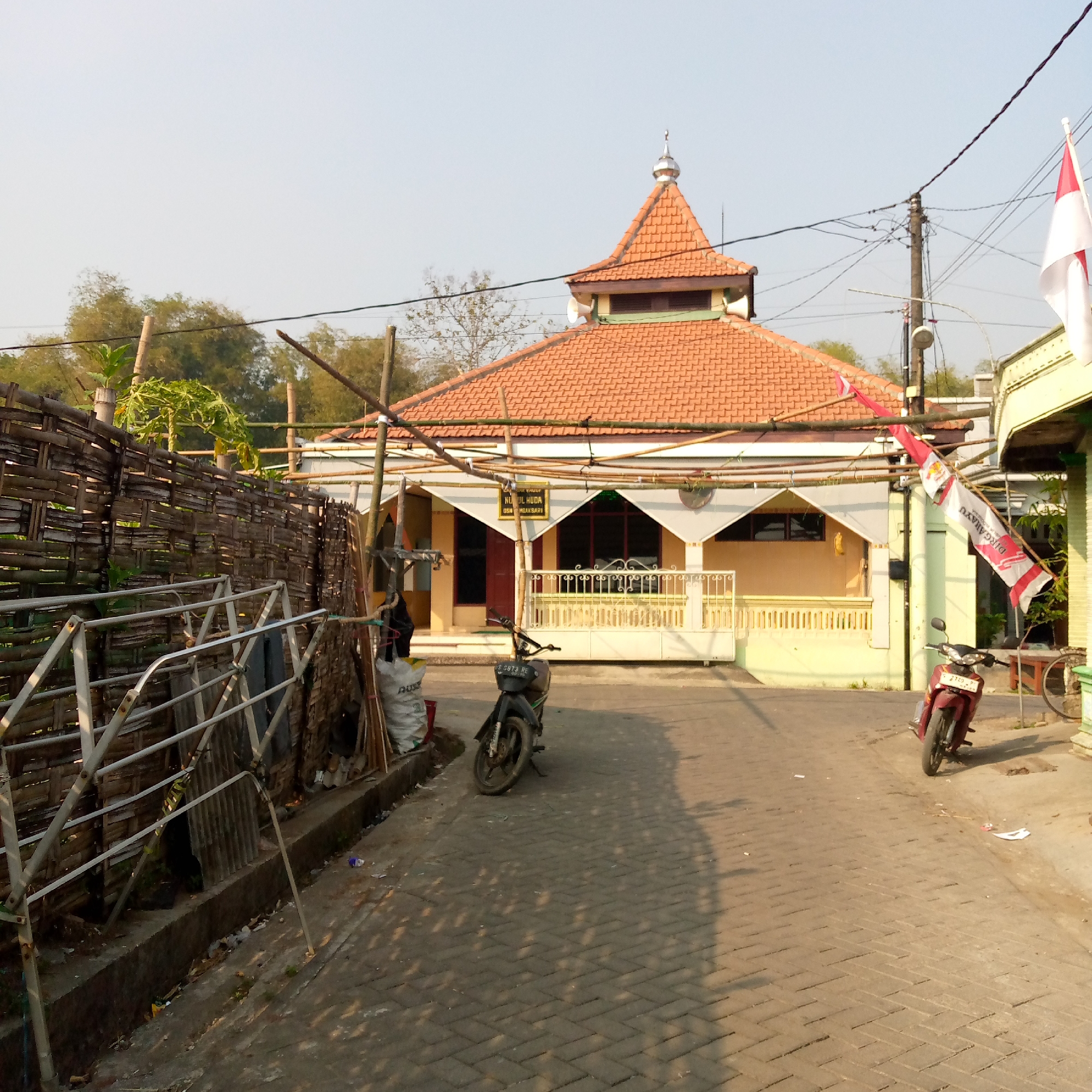A moderate-angle, medium-shot photograph captures the exterior of the "Masjid NURUL HUDA" in Tambakagung, Indonesia. The mosque, characterized by its orange tiled roof and beige-colored walls, is situated on a paved walkway. Two motorcycles are parked on the walkway: a dark-colored one on the left and a maroon one on the right. A bamboo fence borders the left side of the image, and a flag bearing red and white stripes, likely the Indonesian flag, is visible on the right. The scene appears to be a street in a rural or suburban area, with trees and foliage in the background. The sky is clear and a light blue. The lighting suggests it is daytime, possibly late afternoon, given the long shadows cast on the ground. There are no people visible in the image.
