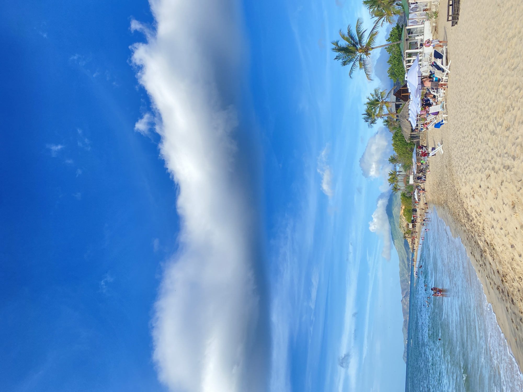 A wide, sandy beach in Altagracia, Venezuela is populated with numerous sunbathers and swimmers enjoying a bright, sunny day. The ocean is a vibrant blue, with gentle waves lapping at the shore. Palm trees dot the landscape behind the beach, and in the distance, rolling hills can be seen under a vast blue sky with scattered white clouds. People are seen relaxing in lounge chairs under umbrellas, walking along the beach, and swimming in the water. Several small structures, possibly beachside cafes or changing rooms, are visible along the shoreline.