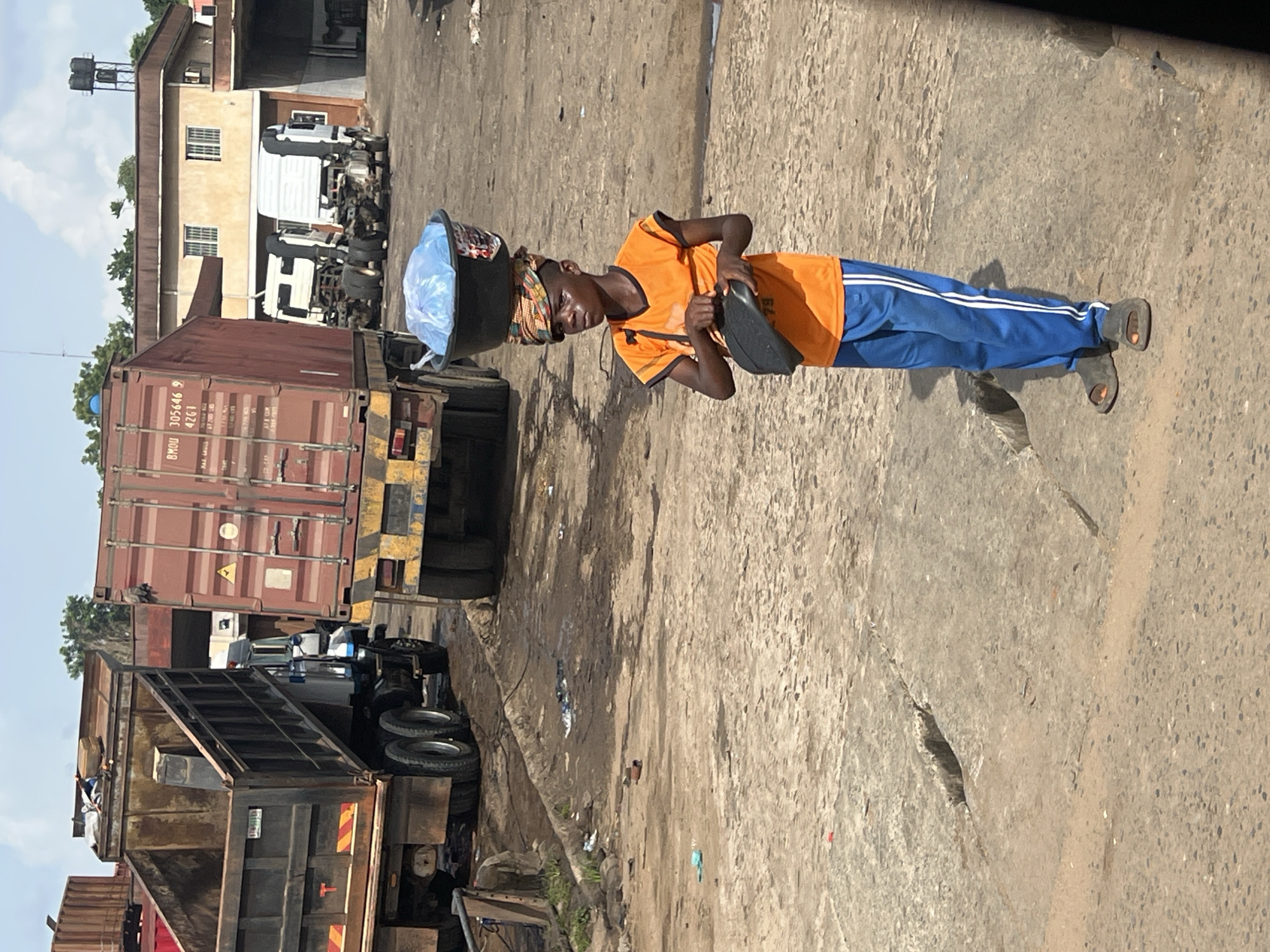The image shows a street scene with several large trucks, possibly a commercial or industrial area. A person is carrying a container on their head while walking along the street. The individual is wearing an orange shirt and blue pants with white stripes. They also appear to be holding something in their hands. The street is made of concrete and has visible wear and tear, with cracks and discoloration. The background includes buildings and other vehicles, suggesting an urban environment. The location is identified as Unknown, Unknown.