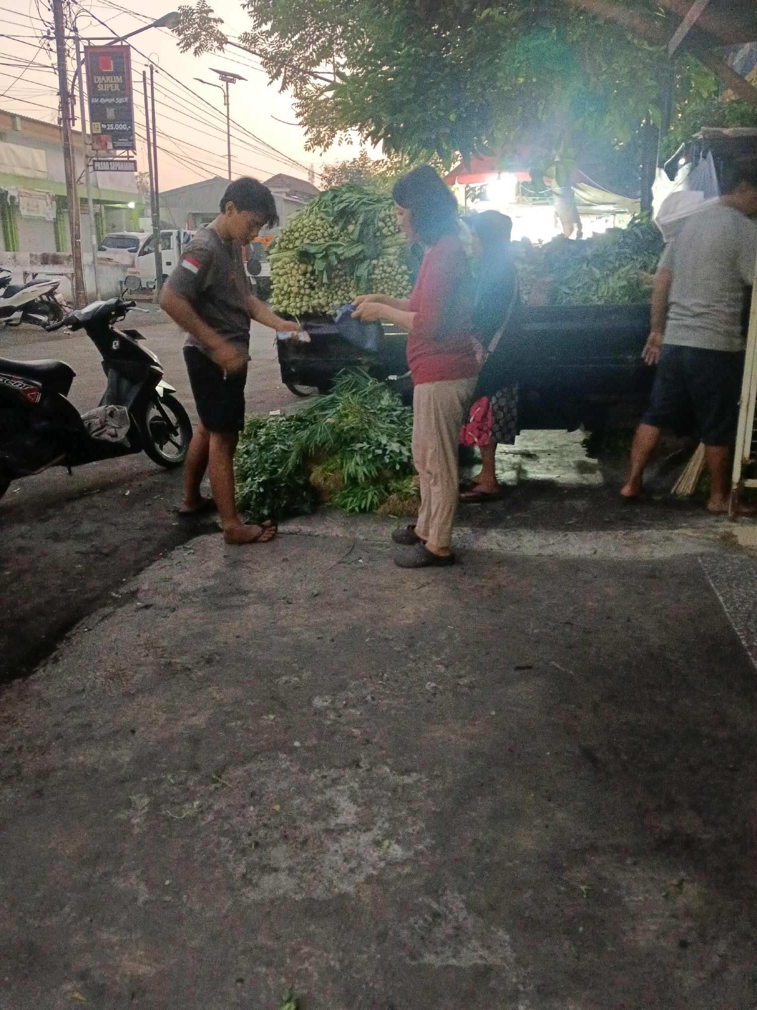This image captures a street scene in Wonocolo, Indonesia, during daylight hours. The foreground shows a concrete ground littered with some debris. In the mid-ground, a young man wearing a grey t-shirt and dark shorts stands next to a large pile of fresh produce, likely vegetables, in the back of a truck. He appears to be interacting with a woman who is wearing a red shirt and light-colored pants. She is holding something in her hands, possibly money or goods. To the left of the man, a black motorcycle is parked on the side of the road. Further back, more people and vehicles are visible, including a white truck and a building with a sign that reads "DJARUM SUPER." The overall environment suggests a bustling marketplace or street vendor area. The lighting indicates it is either early morning or late afternoon, with sunlight filtering through trees.