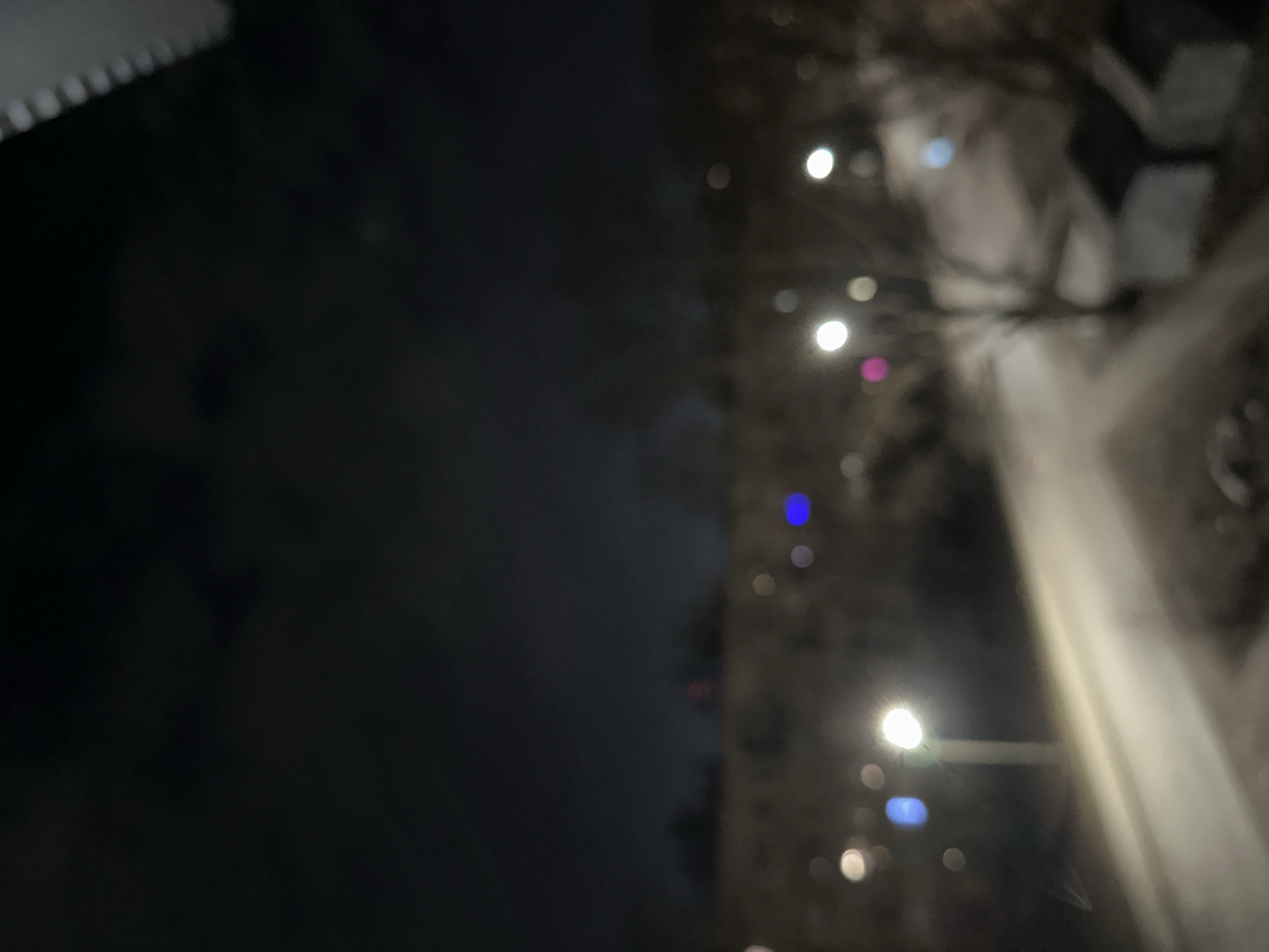 The image appears to depict a street scene at night in an urban environment. Streetlights illuminate the road, casting bright orbs of light and creating streaks on the pavement. Buildings and trees are visible on the right side of the frame, their shapes blurred. The left side of the image is dominated by a dark, indeterminate area, possibly obscured by darkness or atmospheric conditions. The location is Unknown, Unknown.