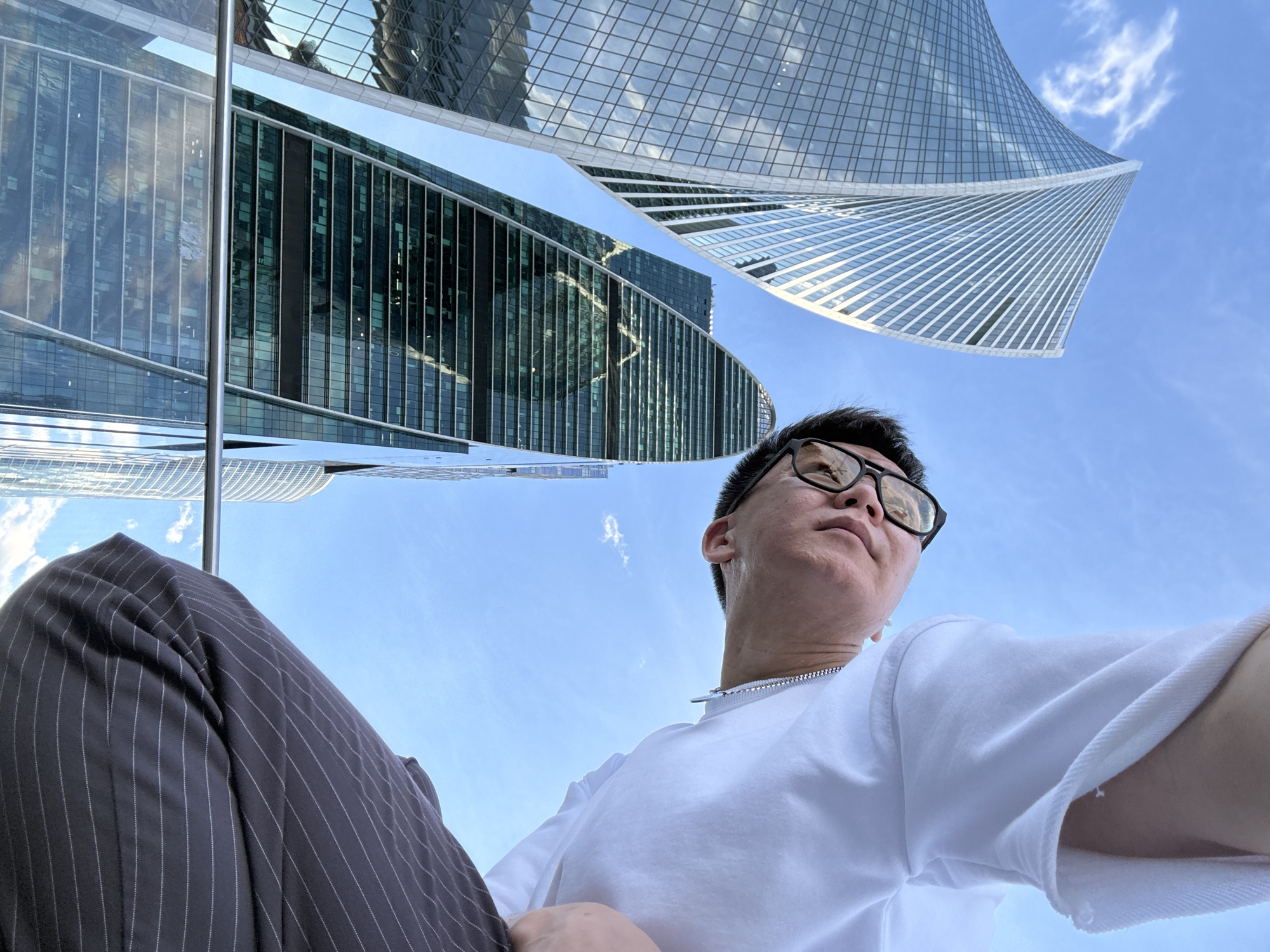 A person is captured in a low-angle, selfie-style shot against a backdrop of modern skyscrapers and a clear blue sky with scattered clouds. The individual, seen from the neck down and with their face partially visible in the upper right quadrant, is wearing a white t-shirt and dark pinstripe pants. They are looking upwards and slightly to the left. The prominent skyscrapers are tall, sleek structures with reflective glass facades, characteristic of a business district. The perspective emphasizes the imposing height of the buildings and the vastness of the sky.