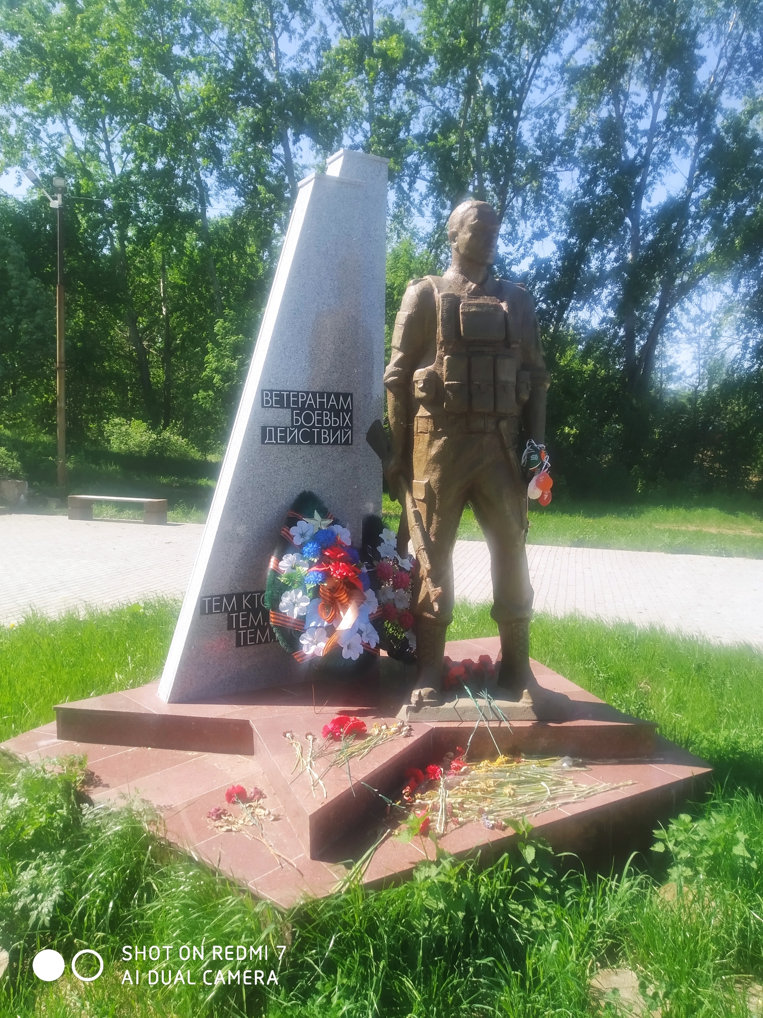 This image shows a monument dedicated to war veterans. The monument consists of a triangular grey stone slab with inscriptions in Russian, a bronze statue of a soldier in combat gear standing next to it, and a red star-shaped base. A wreath of flowers and several red carnations are placed at the base of the monument. The location is Ochansk, Russia. The monument is surrounded by a grassy area and trees. A bench is visible in the background.