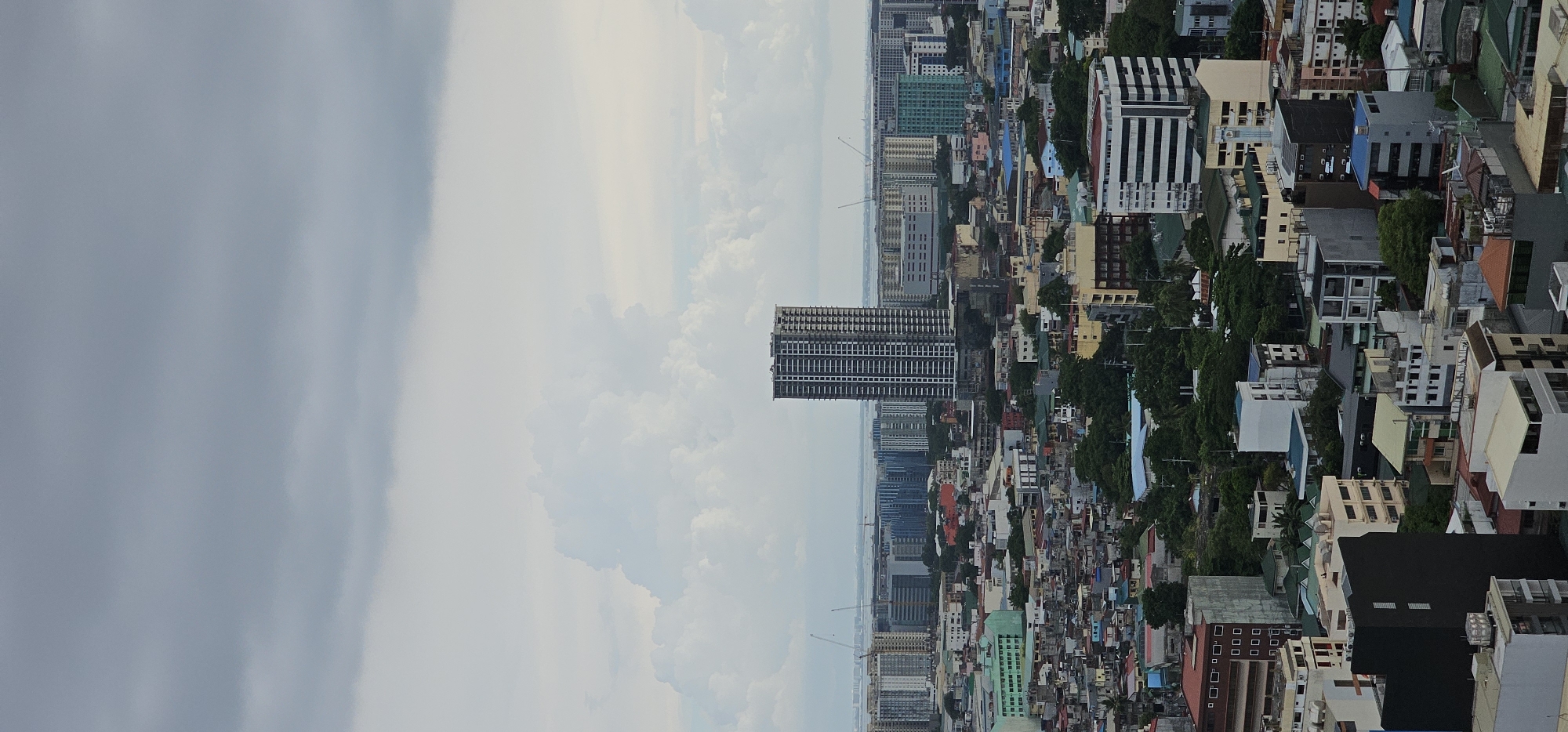 The image depicts an aerial view of a dense urban landscape, likely within the city of Makati, Philippines, characterized by a mix of low-rise buildings, residential areas, and scattered modern high-rise structures. Dominating the skyline is a tall, unfinished skyscraper, its skeletal frame and exposed concrete structure visible against a cloudy sky. In the background, a cluster of contemporary buildings with reflective facades suggests a more developed business district. The foreground and mid-ground are filled with a tapestry of diverse buildings, many with colorful rooftops and varied architectural styles, indicative of a densely populated and established urban area. Greenery, in the form of trees, is interspersed throughout, softening the concrete jungle. The overall atmosphere is one of a bustling city under an overcast sky.