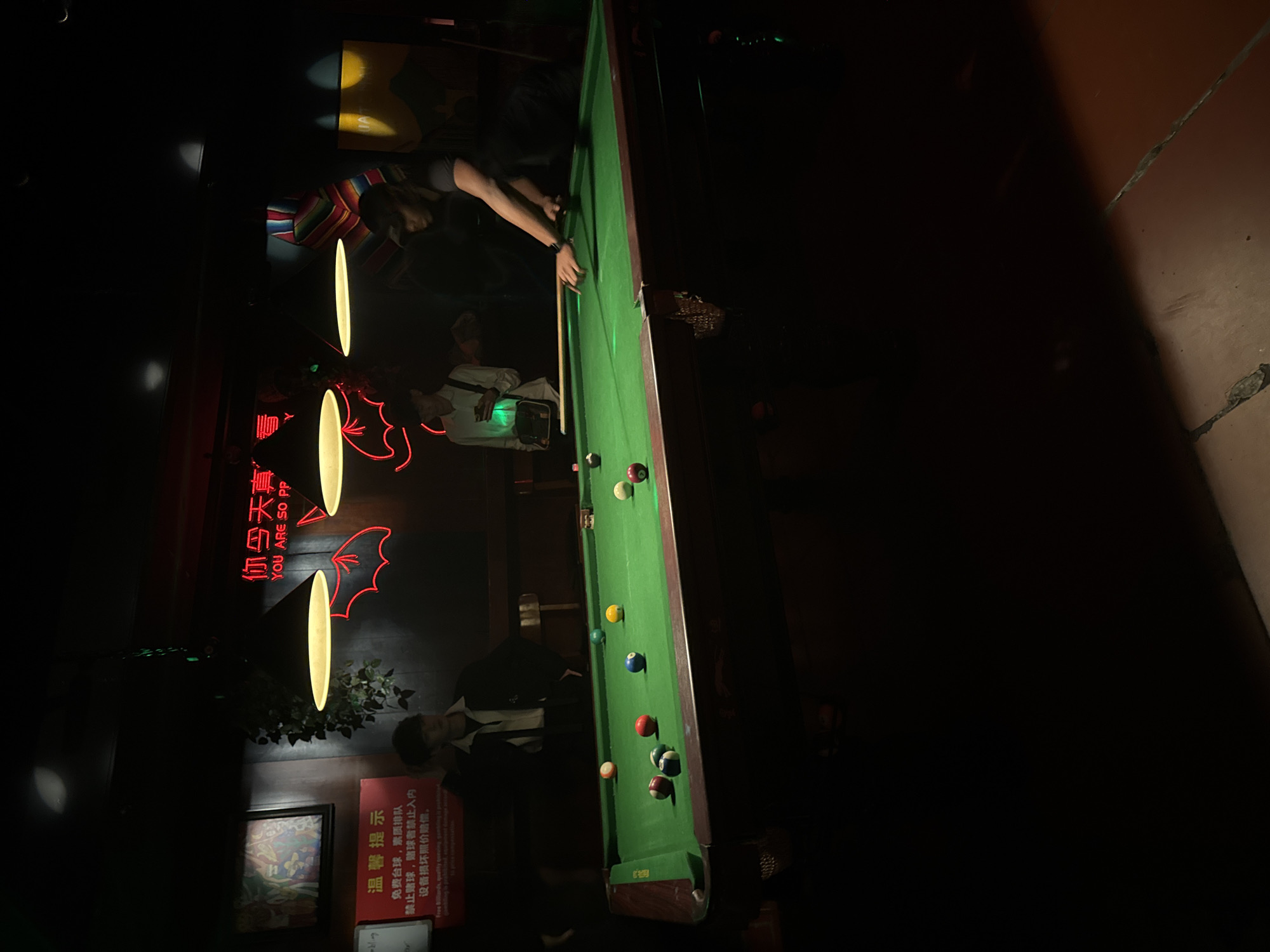 The scene depicts a dimly lit interior of a billiards establishment in Liangjiang, China. A green felt pool table occupies the midground, illuminated by three conical overhead lamps. Approximately 14 multi-colored pool balls are distributed across the table, alongside a cue stick.
Three male individuals are present. On the right side of the table, one male, wearing a dark cap and a dark t-shirt, is poised with a pool cue, indicating an active game. On the left side, another male, dressed in a white shirt and dark jacket, stands observing the table. A third male is partially visible in the deep background, holding an object that emits a green glow.
The surrounding environment features dark walls with decorative elements. A prominent red neon sign displays Chinese characters above "YOU ARE SO PP" and a red bat-wing motif. To the left, a red rectangular sign with white text provides rules and warnings in both Chinese and partially visible English ("Free billiards, quality queuing, queuing is prohibited, equipment damage attention must be compensated"). Other wall decorations include framed artwork, a hanging potted plant, and a striped blanket or textile. The floor consists of brown square tiles.