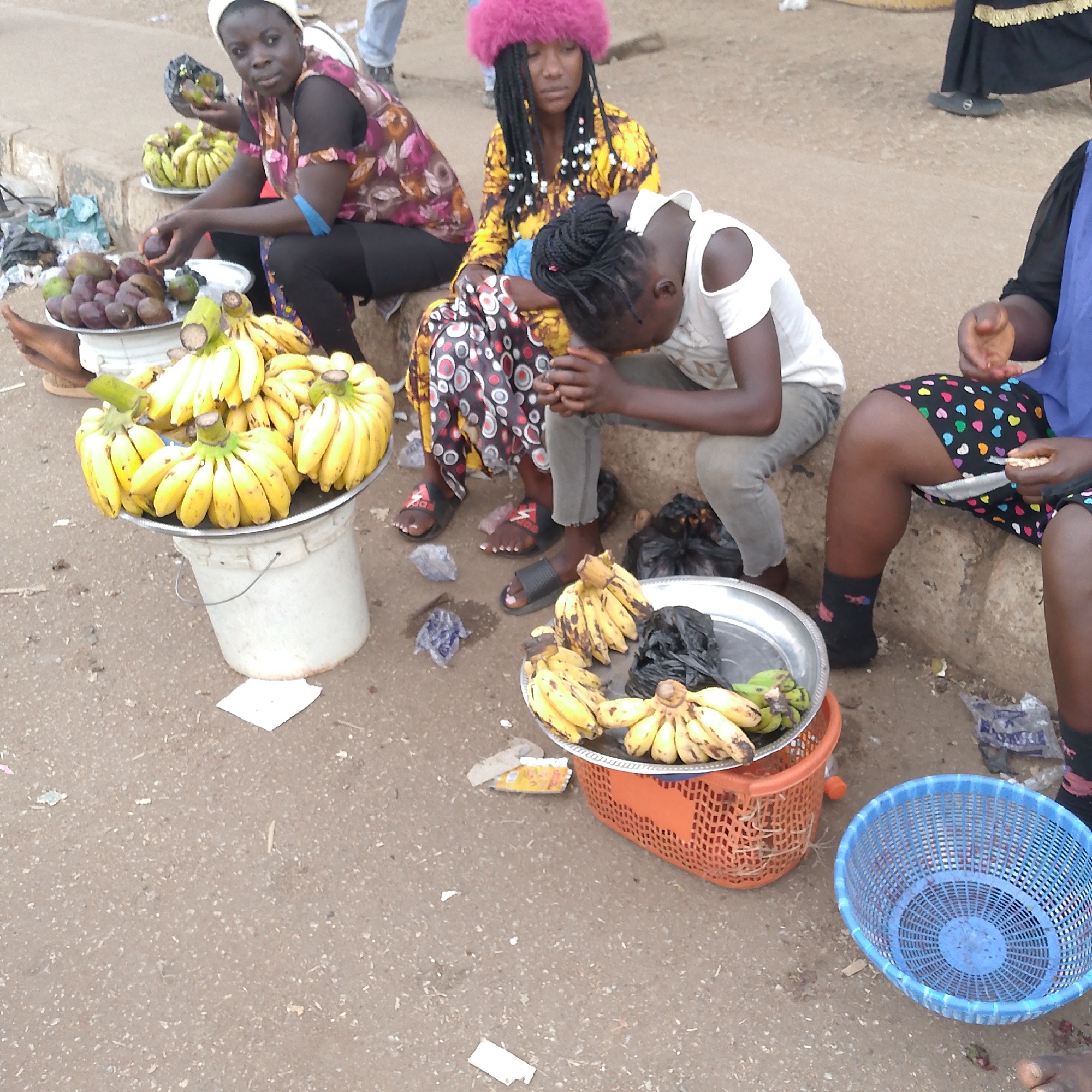 The image depicts a street scene in Bukuru, Nigeria, where several women are selling fruits. One woman, wearing a floral top and black leggings and a white head covering, sits on a concrete ledge, handling avocados and bananas, a tray of purple fruits sits in front of her. Next to her, another woman with a pink fur hat and yellow patterned dress sits on the ledge. A younger woman in a white shirt and grey pants sits near them, looking down with hands clasped. To the right, a woman in a blue top and patterned shorts is also sitting on the ledge, peeling something with her hands. In front of them are containers of bananas, some displayed on a metal tray on a bucket, others on a tray in an orange basket. A blue basket is visible on the ground. The ground is littered with debris.