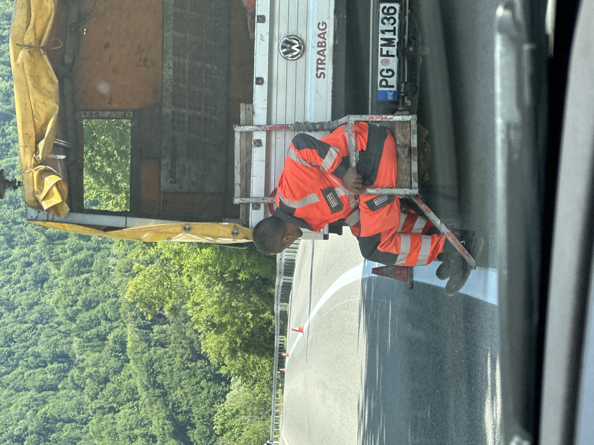 In the city of Crkvine, Montenegro, a man in a high-visibility orange and gray work suit is seated on the rear step of a white Volkswagen truck. The truck has a yellow tarp covering its bed, and the name "STRABAG" is visible on the side. The truck's license plate reads "PG FM136". The scene appears to be on a paved road with a white line being painted down the center. Several orange traffic cones are placed along the side of the road, suggesting ongoing road work. In the background, a dense forest covers a hillside, indicating a rural or mountainous setting. The perspective suggests the photo was taken from another vehicle traveling behind the truck.