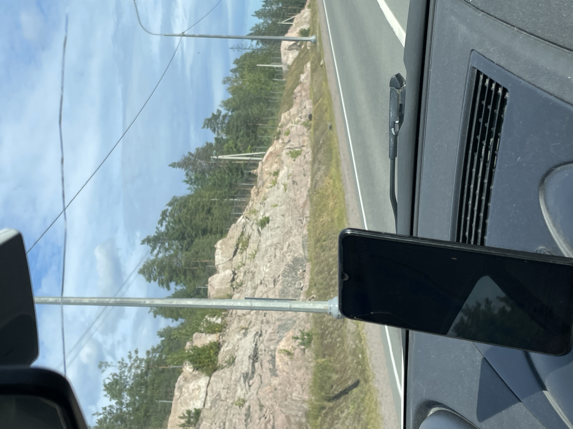 The image shows a view from inside a car, looking out through the windshield. A black smartphone is mounted on the dashboard in the foreground, displaying a reflection of trees and sky. The car is driving on a paved road with white lane markings. To the left of the road, there is a rocky hillside covered with sparse vegetation and pine trees. Overhead, power lines are visible against a blue sky with scattered clouds. The scene appears to be a rural or suburban area, likely on the outskirts of a city. Based on the visual cues, this location could be the city of Kuznechnoye, Russia, which is known for its natural landscapes and proximity to Lake Ladoga.