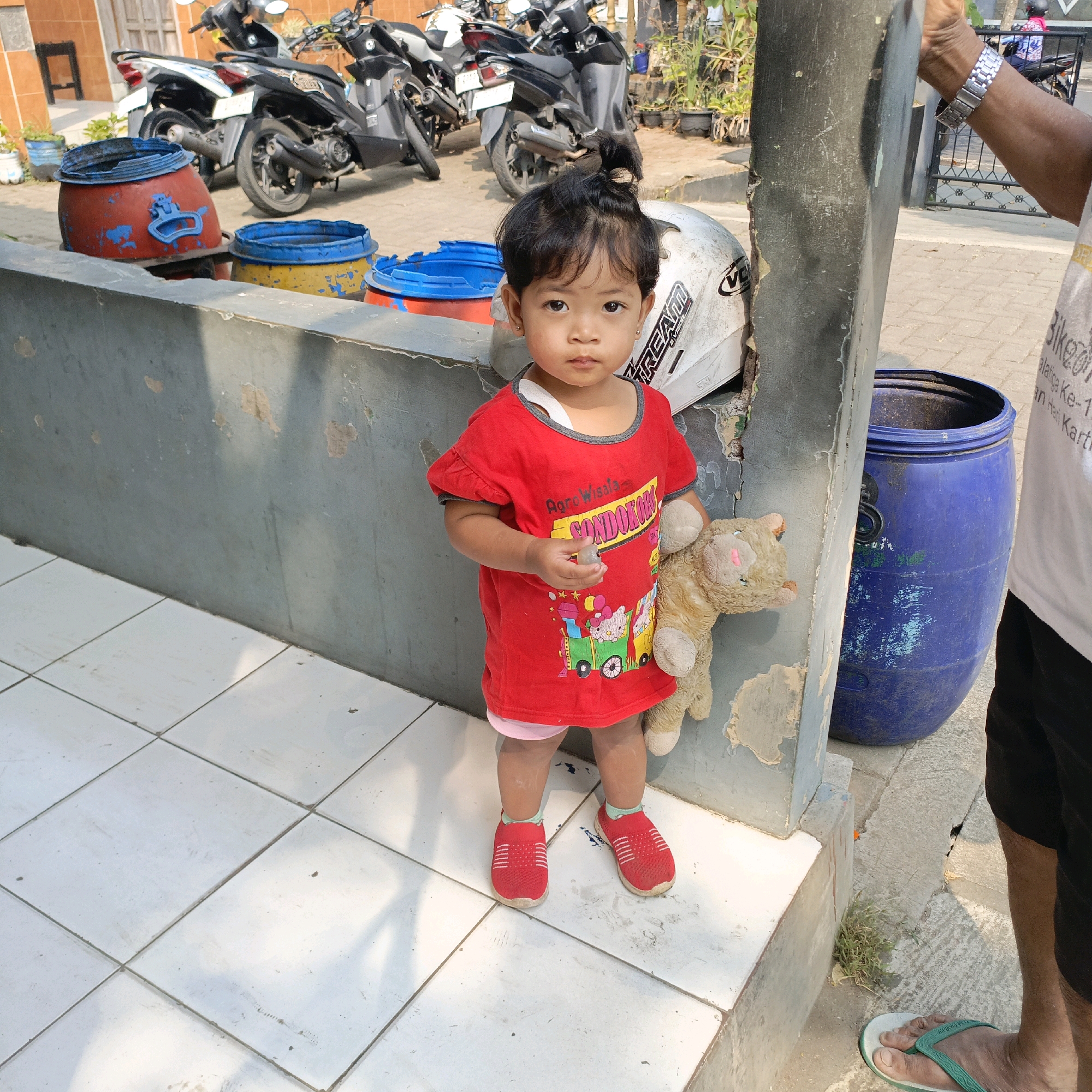 A young child with dark hair pulled into a bun stands on a tiled walkway in front of a concrete wall. The child wears a red t-shirt with a cartoon print and pink shorts, red slip-on shoes, and a light-colored object in their right hand. The child is holding a stuffed animal that resembles a bear. To the left of the child are two large plastic barrels, one red and one yellow with blue rims. Behind the child and barrels are several parked motorcycles. To the right of the child is a large blue plastic barrel. In the background, a person wearing a white t-shirt and black shorts is partially visible, as are more motorcycles and a metal gate. The image appears to be taken outdoors in daylight, with shadows indicating the sun is to the right. Based on the presence of multiple motorcycles and the style of the surrounding buildings and vehicles, the scene is likely located in a residential or urban area in Salatiga, Indonesia.