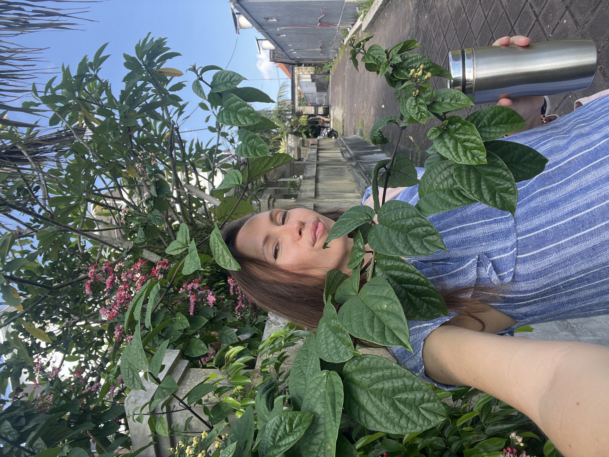 A woman takes a selfie outside in North Kuta, Indonesia. She holds a stainless steel thermos, surrounded by green foliage and pink flowers. The background reveals a narrow alleyway bordered by buildings and a partially visible blue sky. The time of day appears to be daytime.