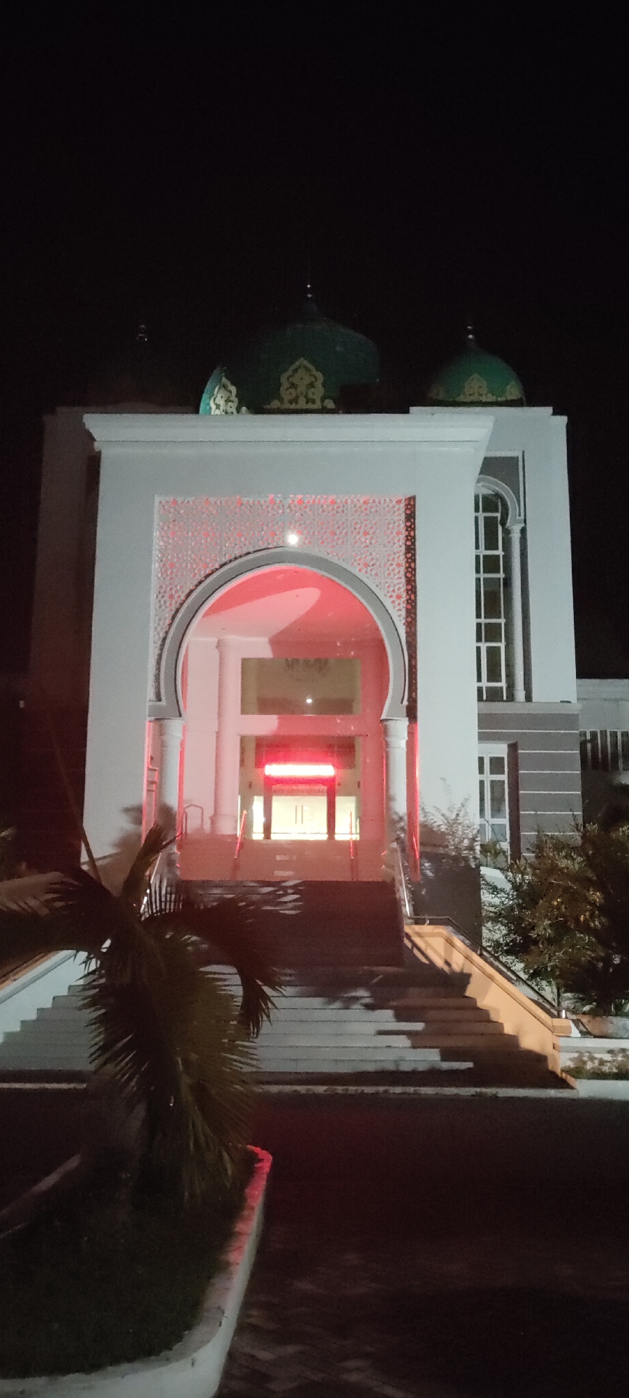 At night, the Baiturrahman Mosque in Lhokseumawe, Indonesia, stands illuminated. The main entrance is framed by a large white archway decorated with a lattice pattern. The arch is lit with red light. Inside, the mosque's interior has additional red lighting. A set of wide steps leads up to the entrance. The structure has green domes. A palm tree is visible in the foreground. Other plants are growing at the base of the stairs. The building has white pillars and windows.