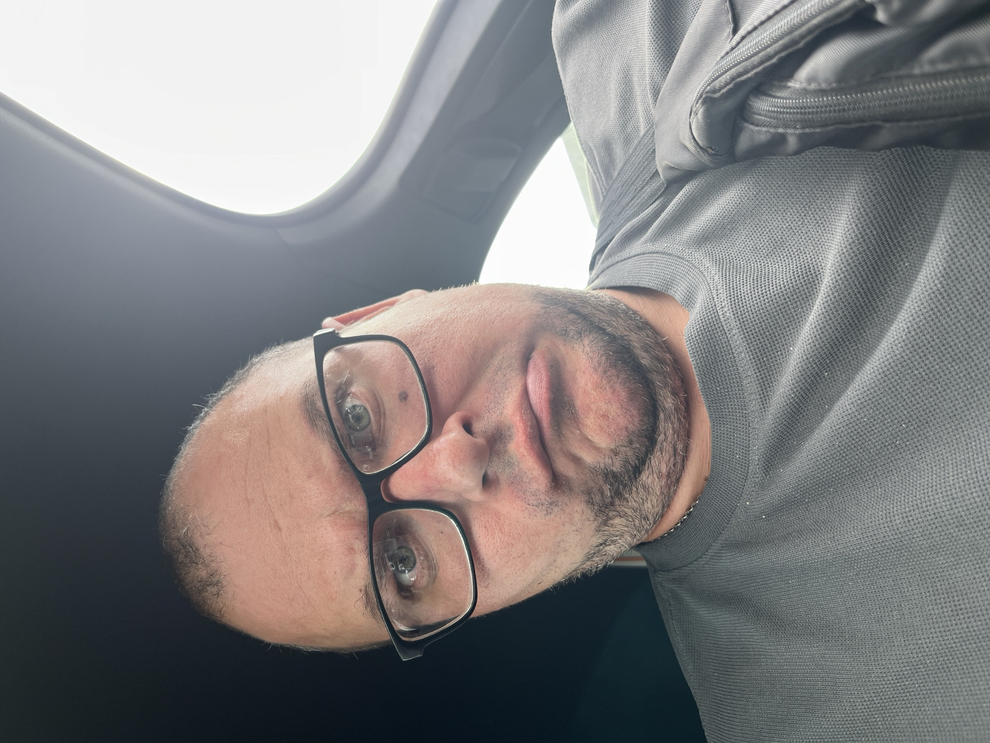 A man wearing black-rimmed glasses and a grey waffle-knit shirt is taking a selfie inside a vehicle. He has a bald head with some sparse hair on the sides and is facing the camera with a grimacing expression. The interior of the vehicle is visible, with a dark headliner and a bright window on the right side, possibly indicating a cloudy day or early morning/late evening light. There is a black bag with a zipper visible on his right shoulder. The image is a close-up, focusing on the man's face and upper torso. There is no identifiable information that places this image in the city of Minsk, Belarus.
