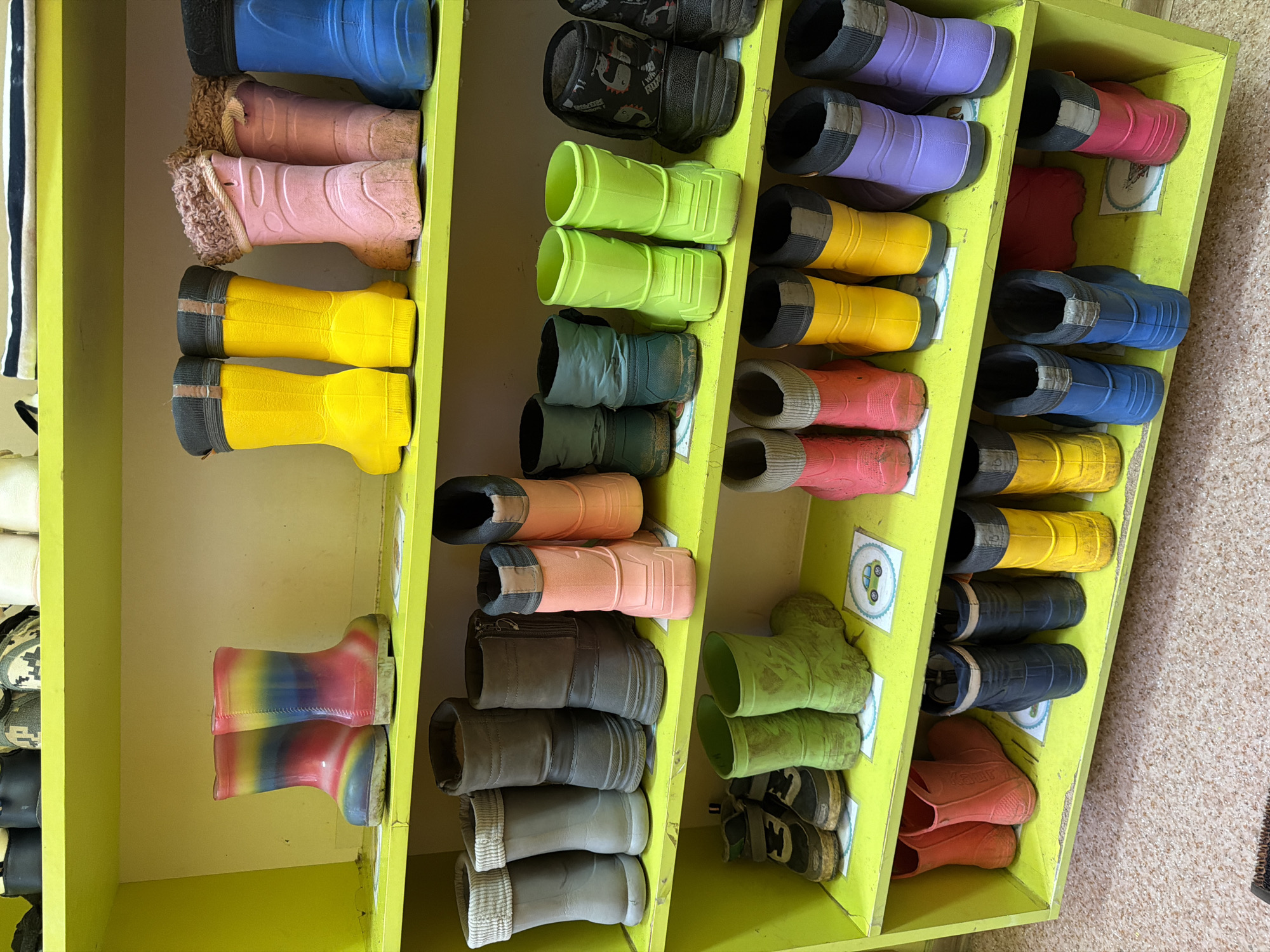 The image shows a yellow wooden shoe rack filled with various pairs of children's boots and shoes. The footwear is arranged in multiple rows, with a variety of colors and styles visible, including blue, pink, yellow, green, purple, and multi-colored options. Some of the boots appear to be made of rubber or a similar waterproof material. The shoe rack appears to be located indoors, likely in a daycare or kindergarten setting. The location of Chaikovsky, Russia is mentioned in connection with this image.