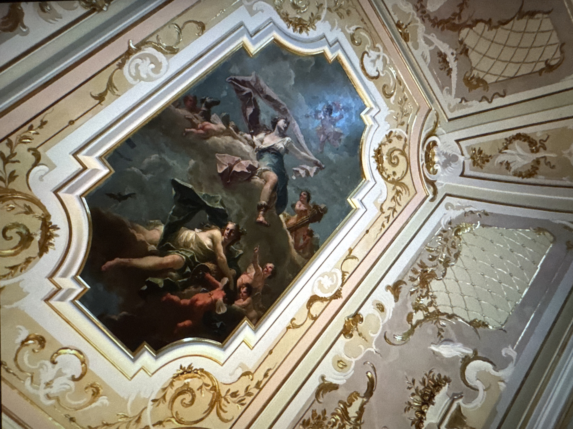 The image shows a frescoed ceiling in a room. The fresco depicts a scene with multiple figures in the sky, rendered in a classical style. The ceiling is ornate, with elaborate stucco work and gilded decorations in gold and cream tones. The architectural details include moldings and decorative panels. The scene in the fresco appears to be mythological or allegorical, with figures in flowing drapery. The overall impression is one of opulence and artistic grandeur. This ceiling is located in the House of the Blackheads in Riga, Latvia.