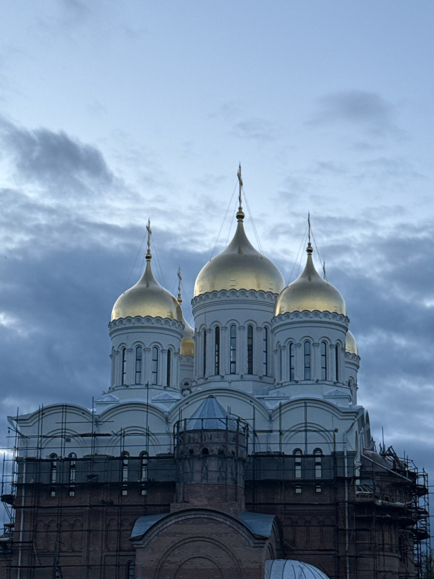 The image shows a large, white Orthodox church with golden domes, under construction in the city of Rusanovo, Russia. The church has multiple domes, with the largest central dome dominating the composition. Scaffolding is erected around the lower brick portion of the building, indicating ongoing renovation or construction work. The sky is overcast with dark, dramatic clouds, creating a moody atmosphere. The scene is a view of the church from a slightly low angle, focusing on its architectural features and the surrounding construction elements. There are no people or animals visible in the image.