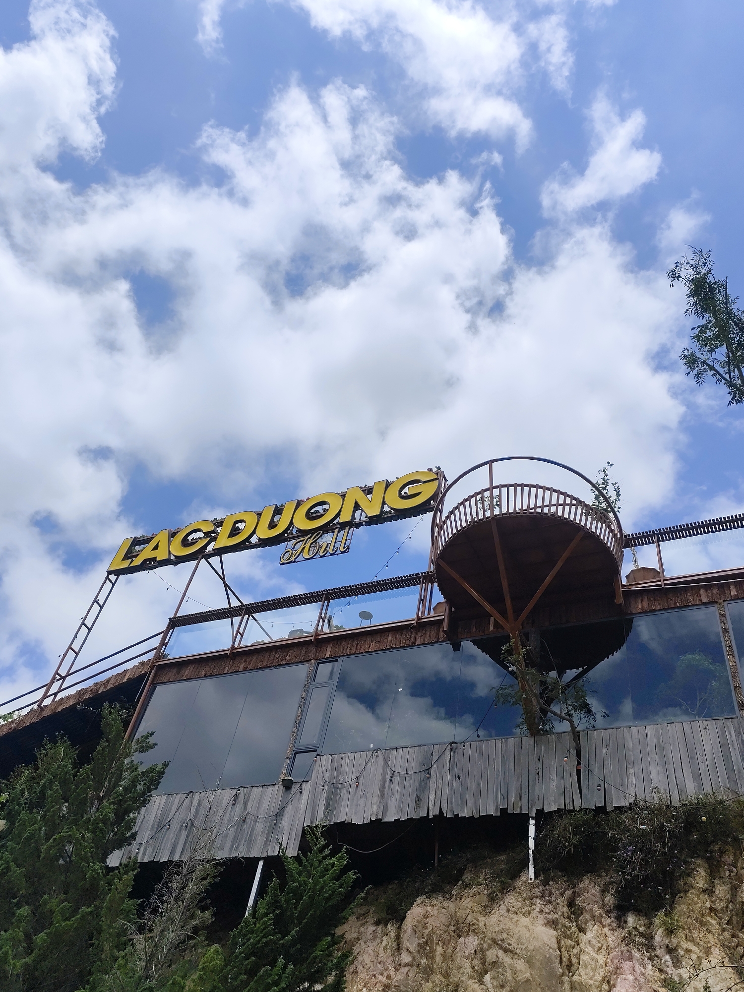 This upward-angled photograph showcases a unique multi-story building, likely a restaurant or cafe, set against a bright blue sky filled with fluffy white cumulus clouds. The structure appears to be integrated into a hillside, with rocky terrain visible at its base, surrounded by green trees and foliage.
Dominating the upper part of the building is a large, illuminated sign with yellow capital letters spelling "LẠC DUONG Hill". To the right of the sign, an unusual circular balcony or observation deck with wooden railings extends outwards, seemingly built around the trunk of a tree that grows upwards through its center.
The building's upper facade features large, dark-tinted glass windows that reflect the sky and clouds, creating an interesting visual effect. Below these windows, the structure transitions to rustic wooden paneling. A ladder-like structure is visible on the left side, leading up towards the sign. Several small string lights are strung along the building's edge.
The time of day is clearly daylight, with abundant natural light highlighting the details of the clouds and the building's materials. No people are visible in the scene, which emphasizes the architectural design and natural setting. The overall impression is one of a distinctive establishment that harmonizes with its natural surroundings.