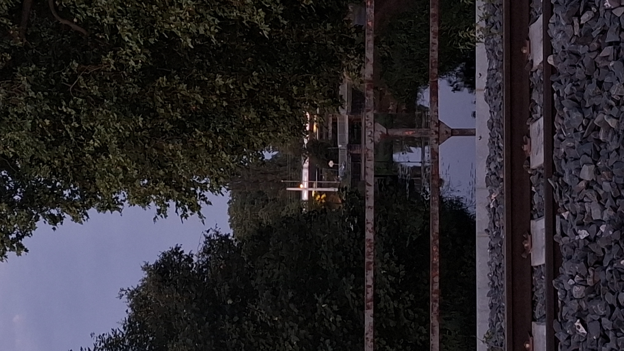 The image depicts a view over train tracks and a rusty railing, looking towards a body of water and some buildings in the distance. Lush green trees frame the scene on both sides. The sky is a dusky blue, suggesting either early morning or late evening. The train tracks are covered in dark grey gravel, with two sets of parallel rails visible. The railing is a weathered, reddish-brown color. The water is dark and reflects the surrounding foliage. In the background, a white building with some illuminated windows is visible, along with what appears to be a tall pole or antenna. The overall impression is of a semi-urban or industrial area with significant natural elements. There are no people or animals visible in the image. The location is not Strasbourg, France.