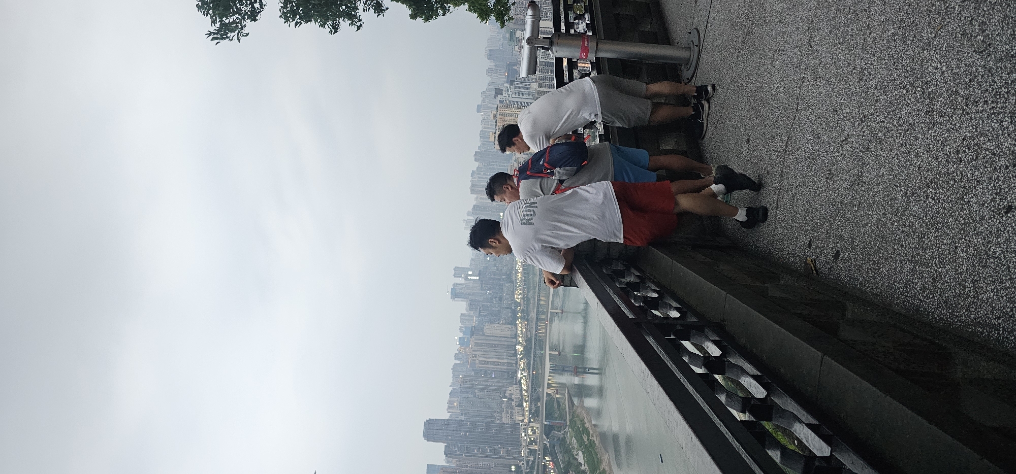 Three men are leaning against a black metal railing, looking out over a city. The man closest to the viewer is wearing a white t-shirt with "RUN" printed on it, and red shorts. The second man is wearing a gray t-shirt, blue shorts, and a blue and red backpack. The third man is wearing a white t-shirt and gray shorts. The cityscape includes many tall buildings and a river running through the center. The sky is overcast. The location is likely in Yuzhong District, China.