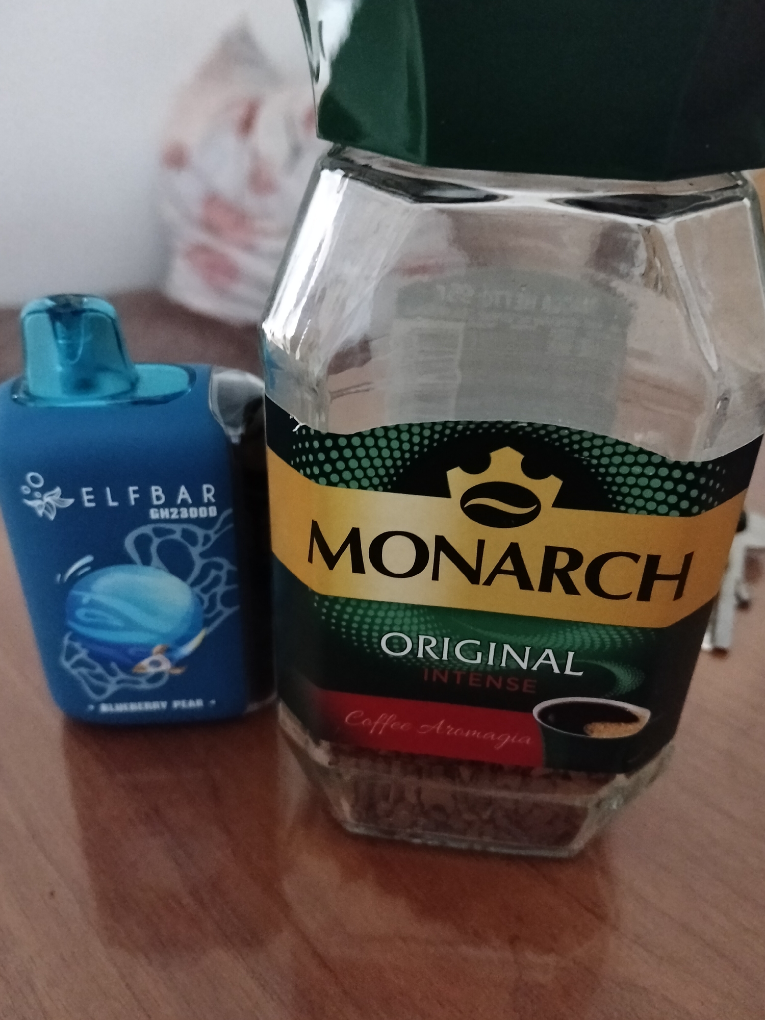 The image shows a close-up of a glass jar of Monarch Original Intense coffee and a blue Elf Bar disposable vape device. The coffee jar is partially filled with coffee grounds and has a green lid. The Elf Bar vape has a blue mouthpiece and is printed with "ELFBAR GH2800B" and an image of a blue orb with lines radiating from it, and "BLUEBERRY PEAR" written below. The objects are placed on a wooden surface, likely a table. In the blurred background, a white plastic bag with red circular patterns is visible. There is no indication of the city of Gorodovikovsk, Russia, or any people in the image. The scene appears to be an indoor setting.