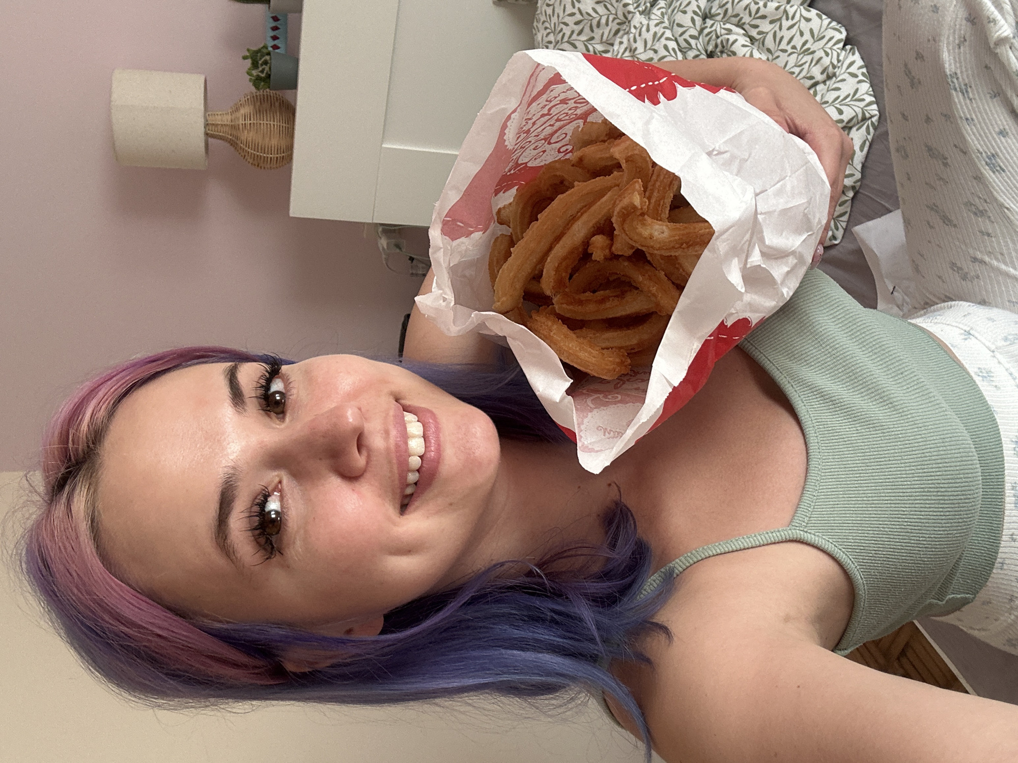 A person with pink and blue hair smiles while holding a white paper bag filled with churros. The person is wearing a light green tank top. A bed with floral patterned sheets is visible in the background. A lamp and bedside table are visible in the top left corner of the image. It is not possible to determine the location in Gijón/Xixón, Spain based on the visual information provided.