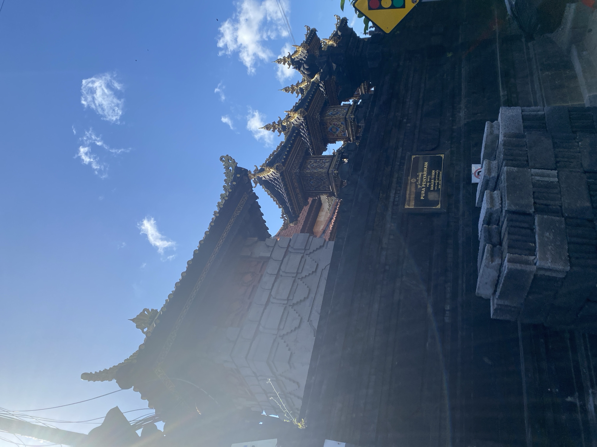 The image shows the Pura Penyarikan temple in North Kuta, Indonesia. The temple is built of dark stone, with a black and gold roof. A small plaque is mounted on the wall, identifying the temple. Stacked stones sit next to the wall. The sky is blue with scattered clouds. A yellow traffic light is visible in the upper corner. Sunlight is shining from the bottom left corner.