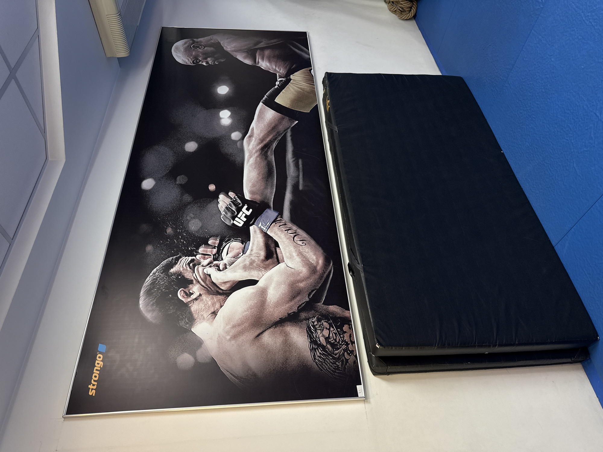 The image shows an indoor setting that appears to be a gym or training facility. On the wall, there is a large poster of a mixed martial arts (MMA) fight scene featuring two male fighters engaged in combat. One fighter is in a dominant position, applying a hold on the other. The poster has a logo "strongo" on the lower left. Next to the poster, there is a folded black mat. The floor is partially white and partially covered with blue mats.
It is not possible to definitively state the location is in the city of Krasnoyarsk, Russia based solely on the image.
