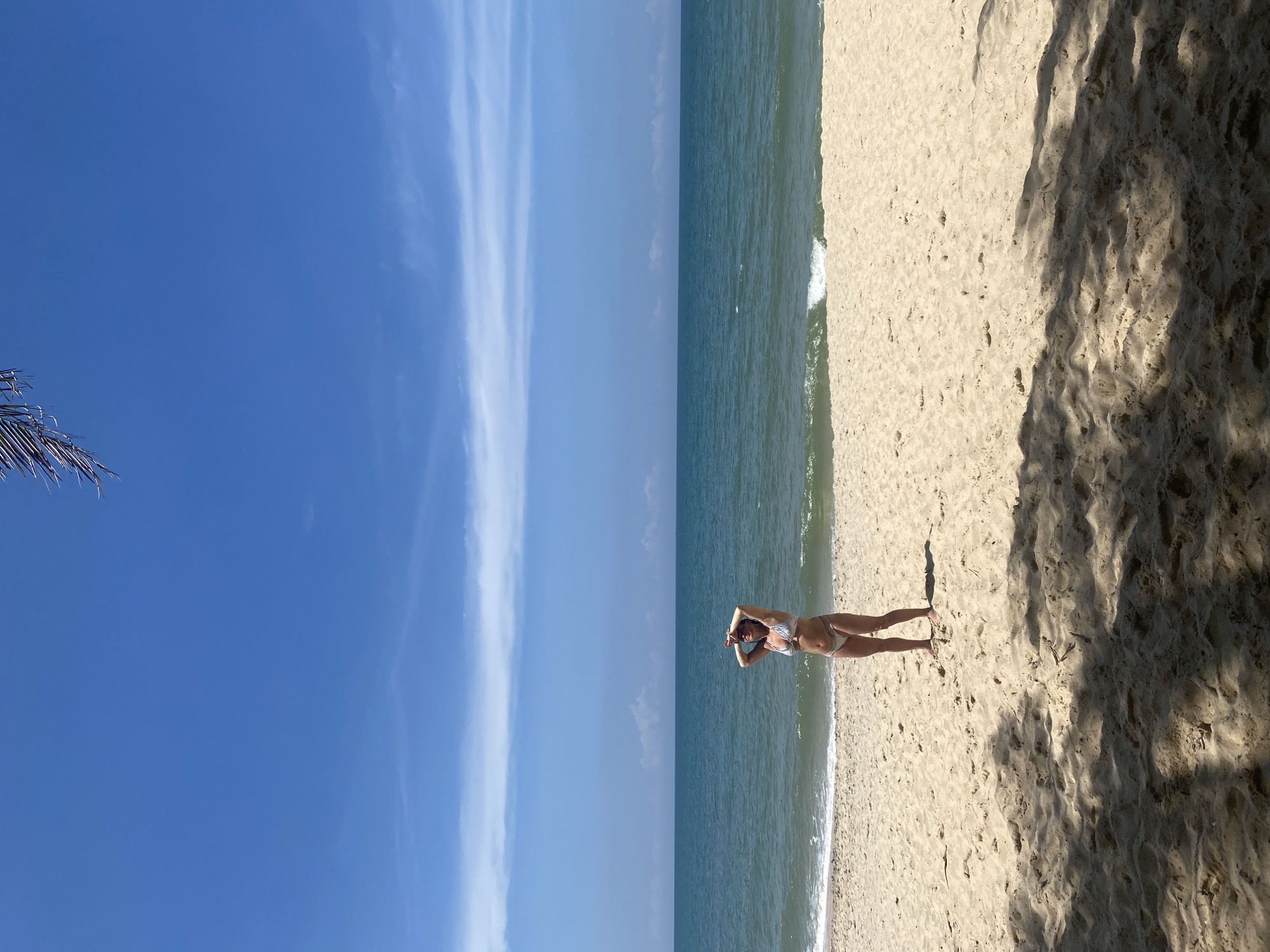 The image depicts a bright, sunny beach scene in Phu Thuy Ward, Vietnam. A single female subject stands on a sandy beach, positioned slightly to the left of the frame's center. She is wearing a light-colored, patterned two-piece bikini. Her arms are raised, and her hands are placed above her forehead, shielding her eyes from the sun. Behind her, the blue-green ocean extends to a clear horizon. A small white wave is breaking on the shoreline to her right. The sky above is a vibrant blue with sparse, elongated white clouds. In the foreground and bottom right, dark, dappled shadows, consistent with overhead foliage such as palm leaves, fall across the textured sand. A dark palm frond is visible in the upper right corner of the frame, partially extending into the blue sky.
