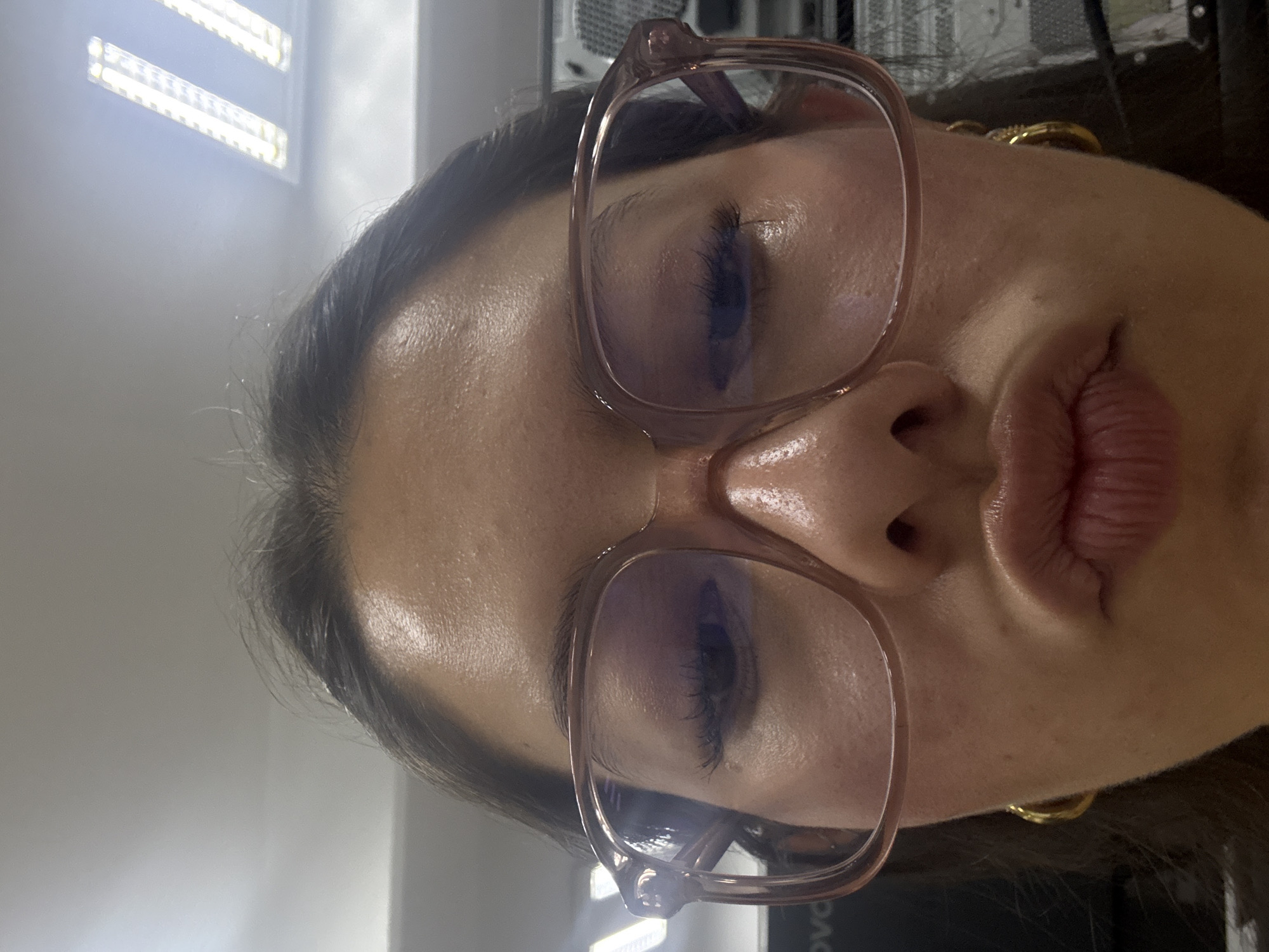 The image is a close-up, sideways portrait of an individual, likely an adult female, captured indoors in San Andrés Cholula, Mexico. The subject's face dominates the frame, appearing to be a selfie. She is wearing eyeglasses with translucent frames, possibly pink or light brown. Her eyes are closed or heavily downcast, showing prominent eyelashes. Her lips are noticeably pursed in a "duck face" expression.
A gold-colored hoop earring is visible on her right earlobe. Her dark hair is swept back or styled away from her face, with some loose strands visible near her forehead. Her skin has a slight sheen, particularly on her forehead and nose, reflecting the ambient light.
The background features bright, rectangular artificial light sources, resembling LED strip lights, suggesting an indoor, artificially lit setting like an office or workshop. Faintly discernible in the blurred background, partially obscured by her glasses and chin, is the text "Lenovo," indicating the presence of a computer or electronic equipment. The overall mood is casual and posed.