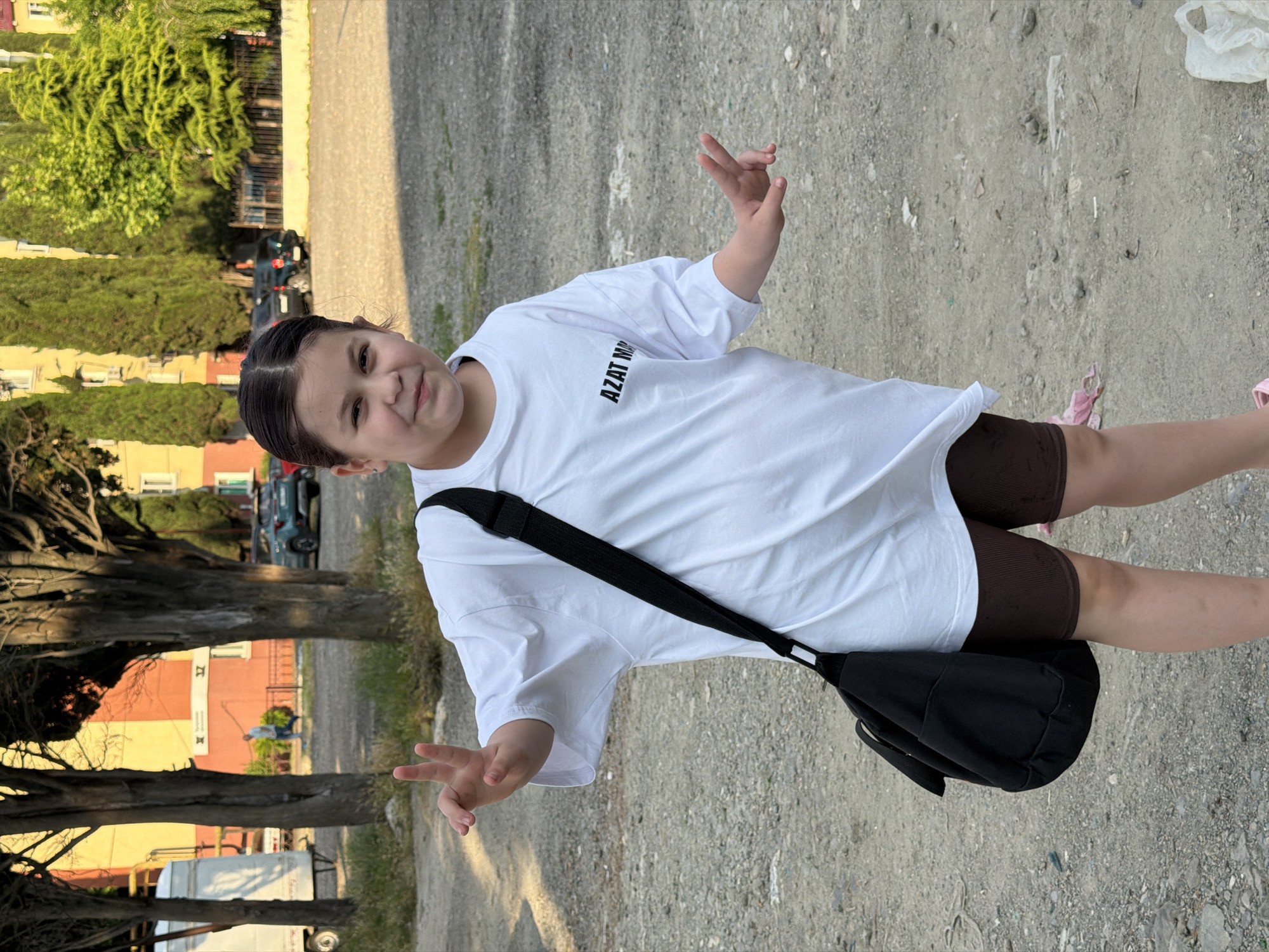 A young girl with dark hair tied back stands outdoors on a gravelly surface, looking towards the camera with a slight smile. She is wearing a white t-shirt with black text that reads "AZAT MM", brown shorts, and has a black messenger bag slung across her body. Her right hand is raised with two fingers extended in a peace sign. Behind her, trees with dark trunks and green foliage line a path. Further in the background, buildings with yellow and orange facades are visible, as well as parked cars and a white van. The lighting suggests it is late afternoon, with the sun casting long shadows. The scene appears to be a residential area or a park.