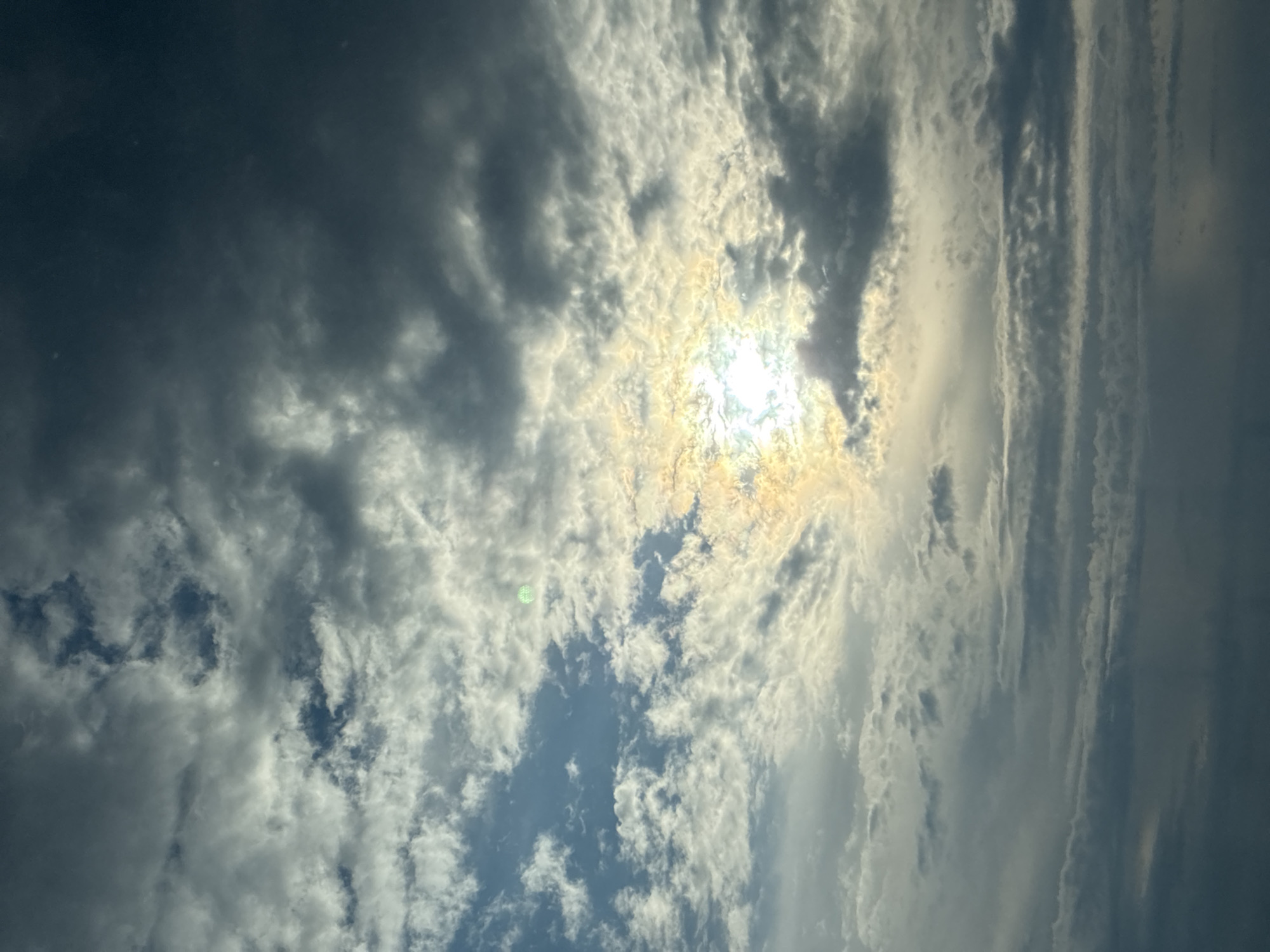An image shows a dramatic sky with a bright sun peeking through dark, ominous clouds. The sun's rays create a halo effect, illuminating the surrounding clouds with a golden glow. The clouds are a mix of dark gray and white, with some appearing wispy and others dense and heavy. The overall impression is one of a dynamic and possibly stormy sky. The image is taken from a low angle, looking up at the sky. There are no visible people or objects in the frame, the location is unknown.