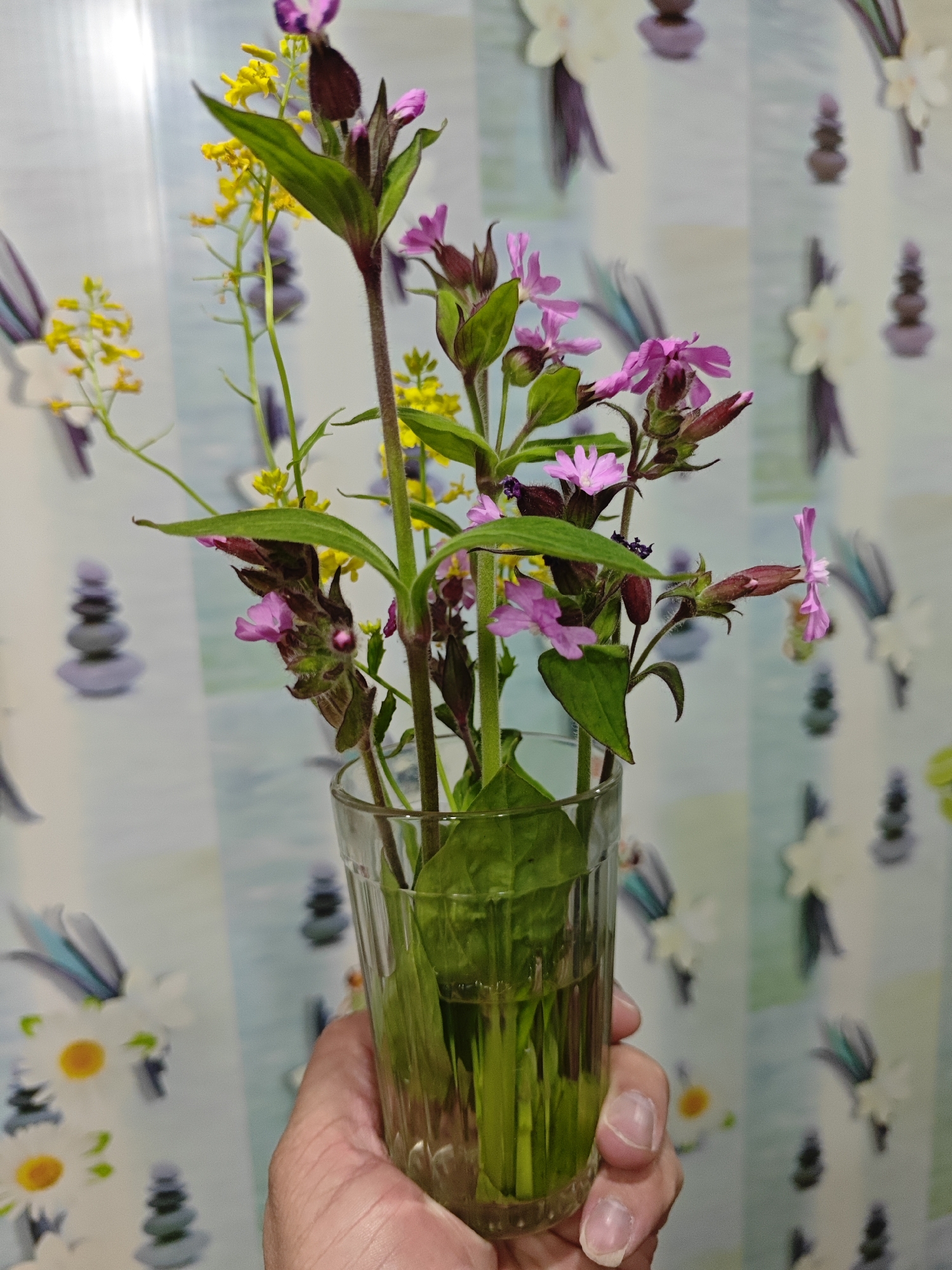 A person's hand holds a clear glass filled with water and a bouquet of wildflowers. The flowers are a mix of pink and yellow, with green leaves. The background is a patterned wallpaper with images of stacked stones and floral designs. The scene appears to be indoors, possibly in a home. There is no indication of the location being in a city.