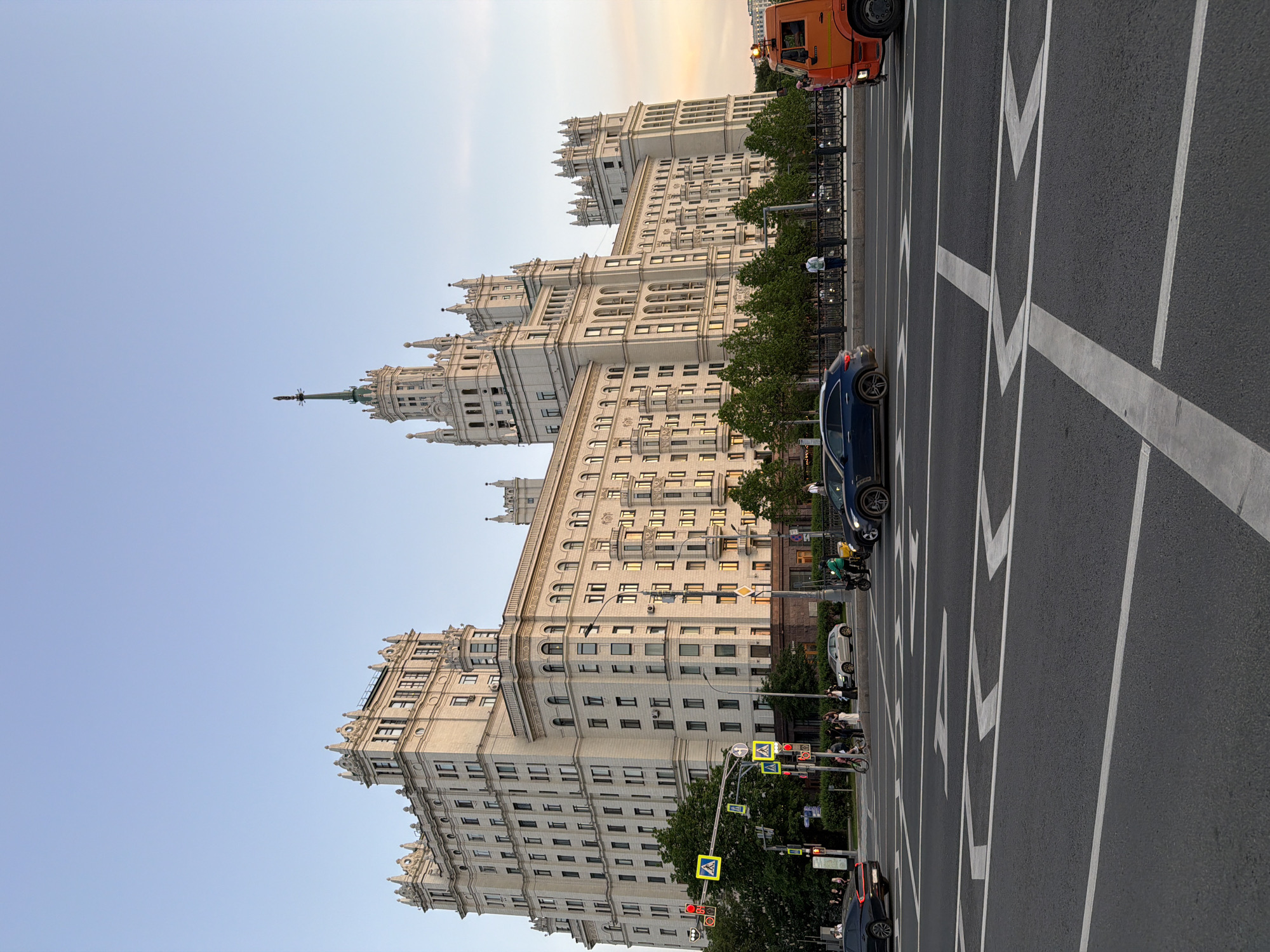 The image captures a street scene in Moscow, Russia. The dominant feature is one of the Seven Sisters skyscrapers, characterized by its Stalinist architecture, including its ornate facade and spire. The building is a light, creamy color and towers over the street.
The street itself is a wide avenue with multiple lanes marked by white chevrons. Several vehicles, including cars, are visible on the road. Traffic lights and pedestrian crossings can be seen, indicating a busy urban environment. Trees line the street, adding a touch of greenery to the scene. The sky is clear and a pale blue.