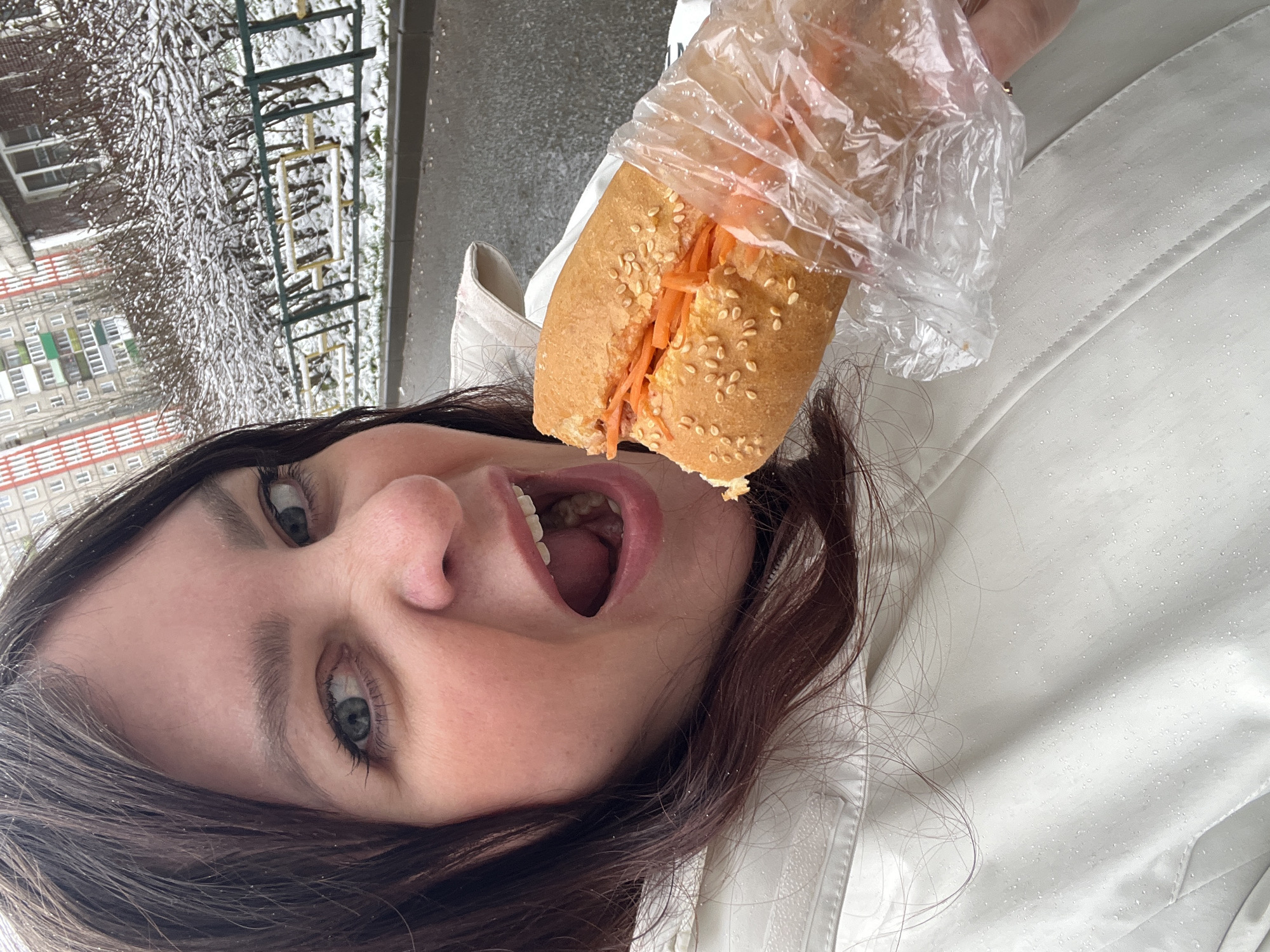 A woman is holding a sandwich in a plastic bag near her open mouth. She is wearing a white jacket and has brown hair and light eyes. The sandwich appears to be a banh mi, filled with shredded carrots and sesame seeds on the bread. Snow-covered trees, a fence, and a multi-story building are visible in the background. The location is Perm, Russia.