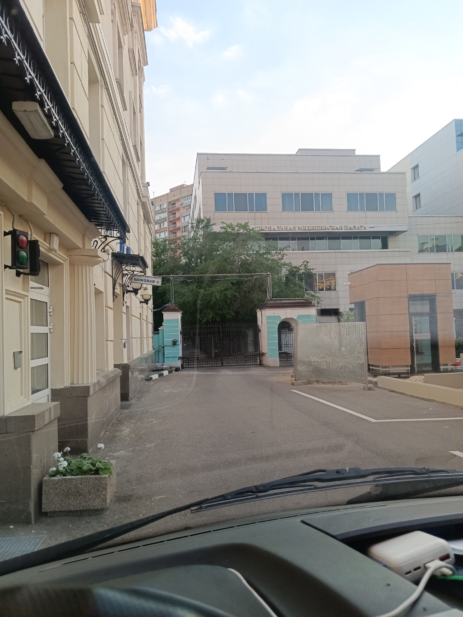 This image depicts an outdoor urban scene in Moscow, Russia, specifically at Pereulok Shokova, 2, identifiable by the visible text "ШОКОВАЯ 2" on a street sign and "НАЦИОНАЛЬНАЯ МЕДИЦИНСКАЯ ПАЛАТА" (National Medical Chamber) on the main building in the background.
The perspective is from inside a vehicle, looking forward, with the dark dashboard and a windshield wiper visible across the bottom of the frame.
On the left, a multi-story, cream-colored building with classical architectural details, including pilasters and decorative black metal awnings, runs parallel to the viewer. A functional traffic light, illuminated with both red and green, is mounted on this building's facade. A concrete planter containing white flowering plants sits at the base of this building.
In the midground, an asphalt driveway or access road extends towards the background. A white parking line is painted on the right side of this asphalt surface. To the left of the driveway, a decorative, light blue and white arched gateway with a black metal fence provides an entrance. To the right of the gateway, a modern, brown-paneled structure with glass elements stands.
The background is dominated by a large, multi-story, contemporary building with a light-toned facade and multiple rows of rectangular windows, clearly marked with the Russian text "НАЦИОНАЛЬНАЯ МЕДИЦИНСКАЯ ПАЛАТА." Further in the distance, another multi-story residential building with balconies is partially visible. The sky overhead is partially cloudy. No people are present in the scene.