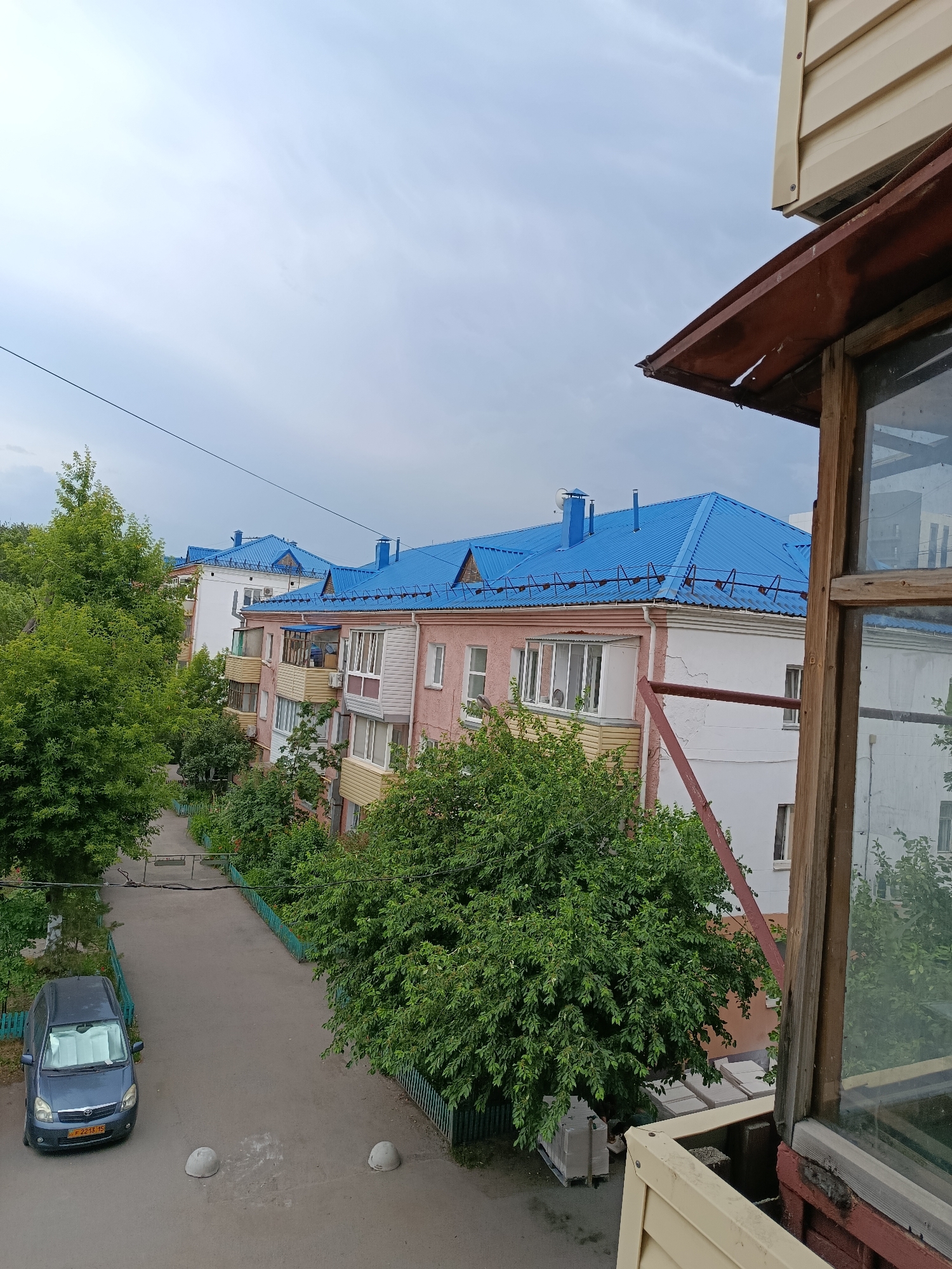 This is an eye-level, medium shot of an urban residential area, likely in Petropavl, Kazakhstan. The scene is dominated by two apartment buildings with bright blue roofs. The building in the foreground is pink and white, with balconies visible. Behind it, another building, mostly white, also features a blue roof. A large, leafy green tree is prominent on the left side of the image, partially obscuring the buildings. In the lower left quadrant, a dark blue minivan is parked on a paved road. The road is bordered by green fences and more trees. In the upper right, a section of a weathered wooden balcony frame with a window is visible, suggesting the photo was taken from another balcony or window. The sky is overcast with pale, wispy clouds. There are no people visible in the image. The scene depicts a quiet, everyday residential street.