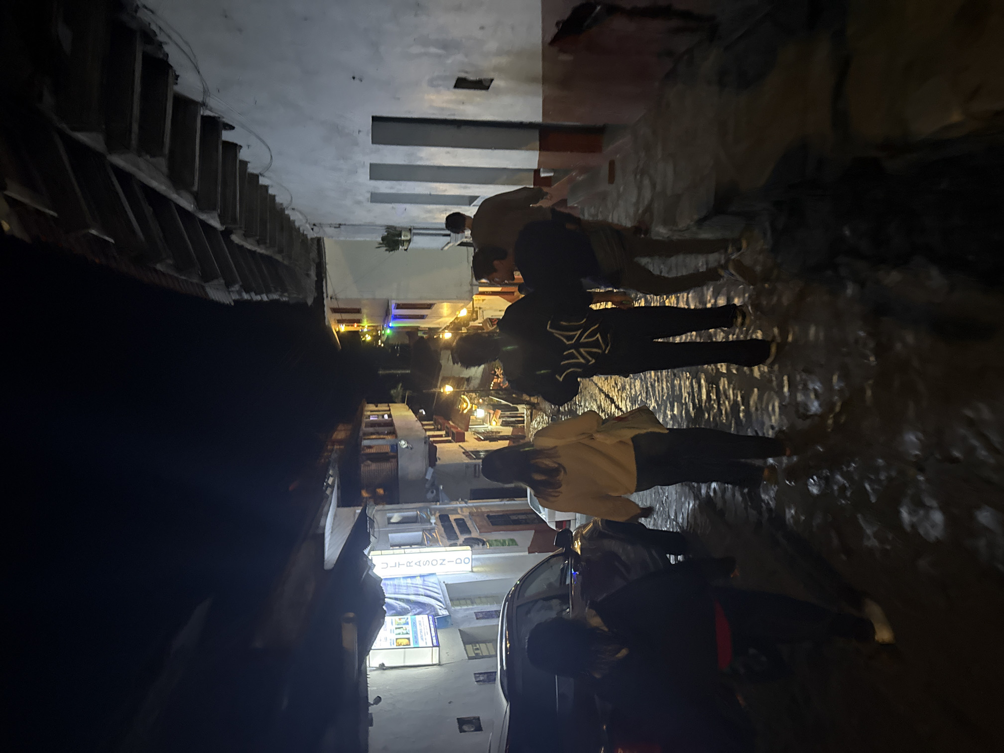An eye-level photograph depicts a dimly lit, narrow alleyway in Cuetzalan, Mexico, at night. Several people are walking along the cobblestone street, their figures partially obscured by shadows. The building on the left has a tiered structure, with visible steps and balconies. On the right, a building shows a pattern of alternating light and dark vertical stripes on its upper level. Streetlights and decorative lights illuminate the scene with a warm glow. A parked car, partially visible, is in the foreground. The atmosphere suggests a bustling, yet intimate urban scene.