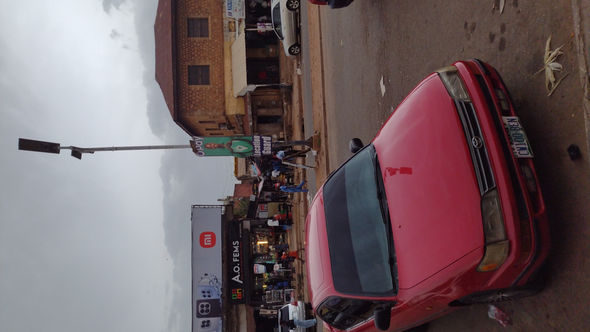 The image captures a street scene in Jos, Nigeria, showcasing a red Toyota car parked on the right side of the road with a "CW100-BEN" license plate. In the background, there's a two-story building with visible windows and stone facade. A street lamp stands tall with advertisements for "Opay" and "Location Guard." Adjacent to the lamp, there's a large advertisement for A.O. FEMS, a point-of-sale (POS) service provider, with a phone number (18206 6246) displayed. The street is bustling with people, suggesting an active urban environment. The sky is overcast, indicating a cloudy or possibly rainy day. A white car is visible behind the red one.