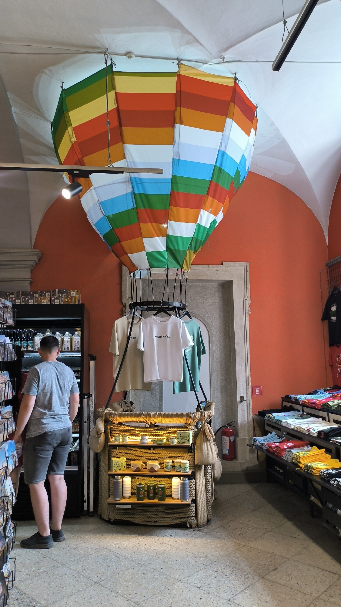 A man in a grey t-shirt and shorts stands in a shop in Lviv, Ukraine, facing away from the camera. In front of him is a display stand resembling a hot air balloon basket, decorated with ropes and burlap bags. The basket holds shelves with various items, including mugs, small decorative items, water bottles, and jars. Hanging above the basket are three t-shirts on hangers: a beige t-shirt on the left, a white t-shirt with text in the center, and a green t-shirt on the right. The hot air balloon itself, a large colorful fabric structure with stripes of green, yellow, orange, red, white, and blue, is suspended from the ceiling. To the left of the display, a refrigerated display case filled with bottles is visible. To the right of the display, shelves are stocked with folded t-shirts in various colors, and a fire extinguisher is mounted on the orange wall. The floor is a mosaic of light-colored tiles. The walls are painted orange.